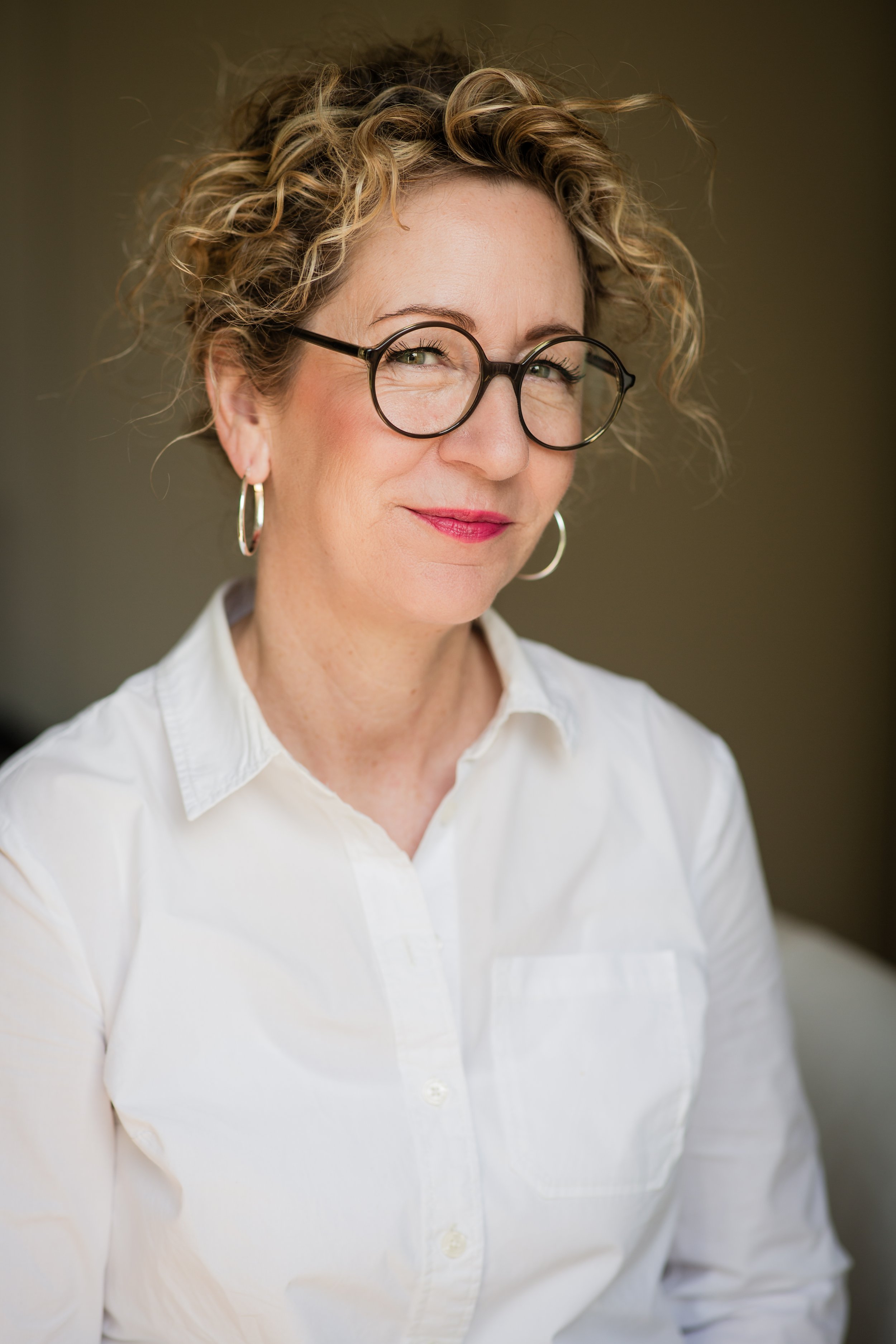 Nahaufnahme einer Frau mit Locken, Brille und silbernen Hoop-Ohrringen, trägt ein weißes Hemd, lächelt leicht, vor unscharfem dunklem Hintergrund.