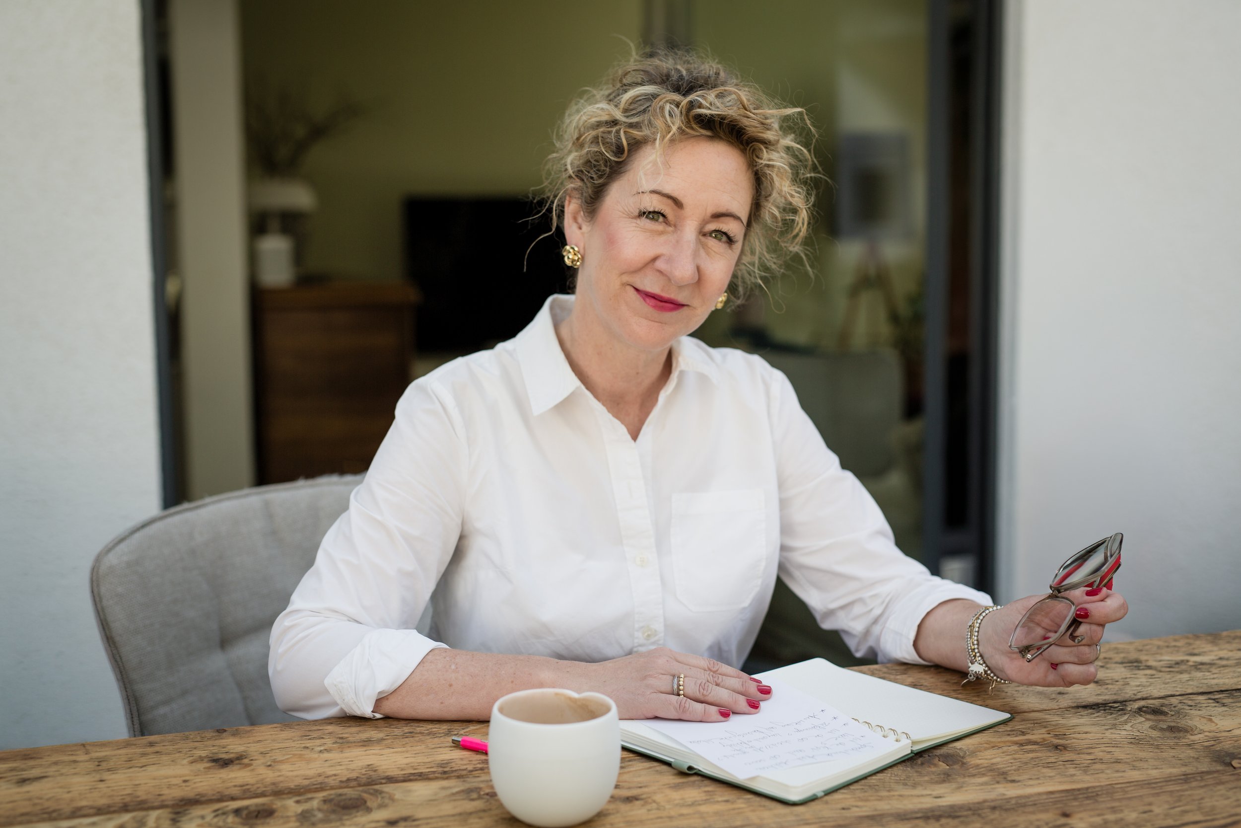 Eine Frau mit lockigem Haar und weißen Hemd sitzt an einem Holztisch, hält ein Smartphone in der Hand, hat ein Notizbuch vor sich und eine Tasse Kaffee darauf. Sie schaut in die Kamera.