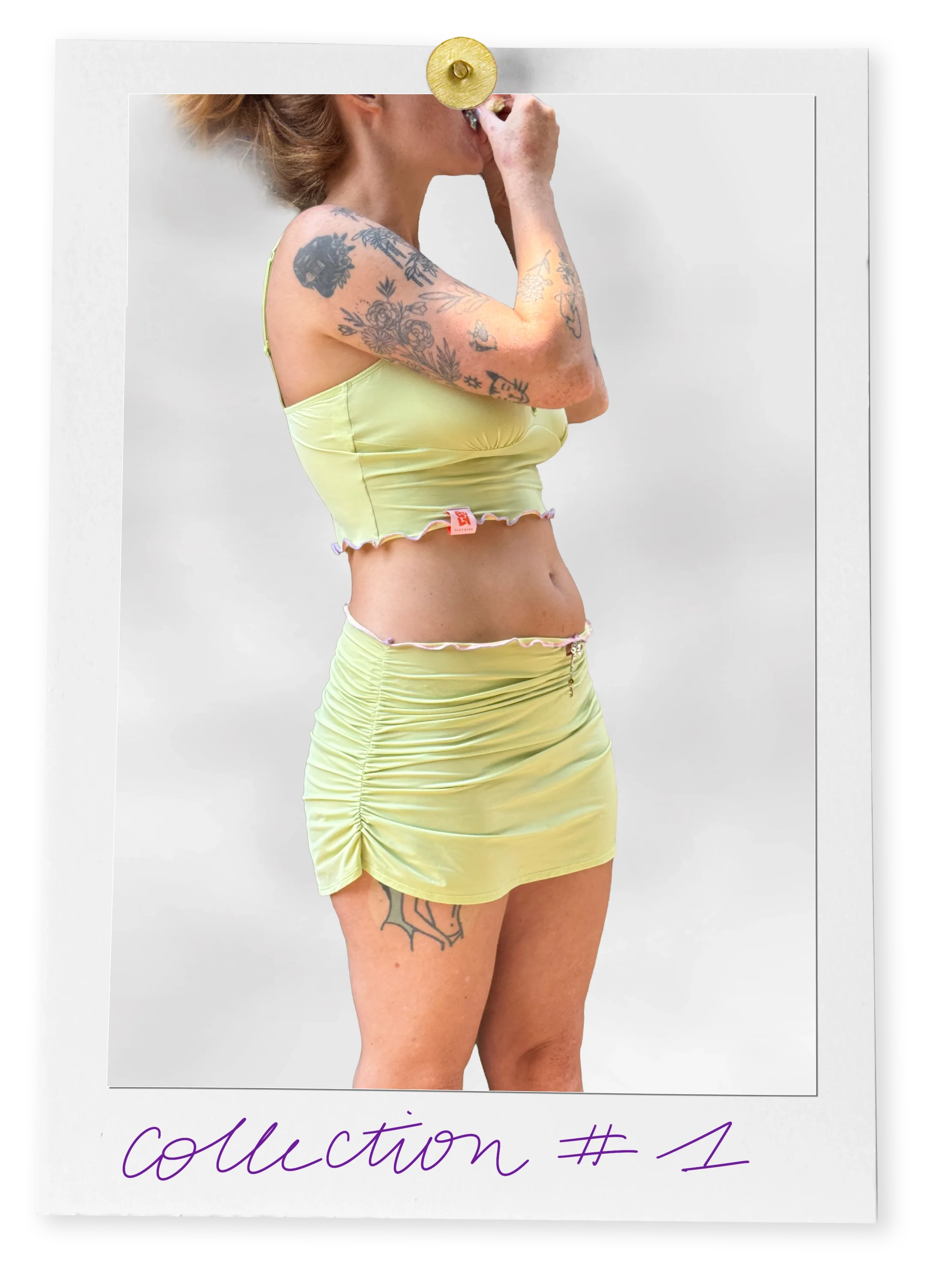 Une femme en vêtements verts, montrant ses tatouages, tenant sa main près de sa bouche sur un fond blanc.