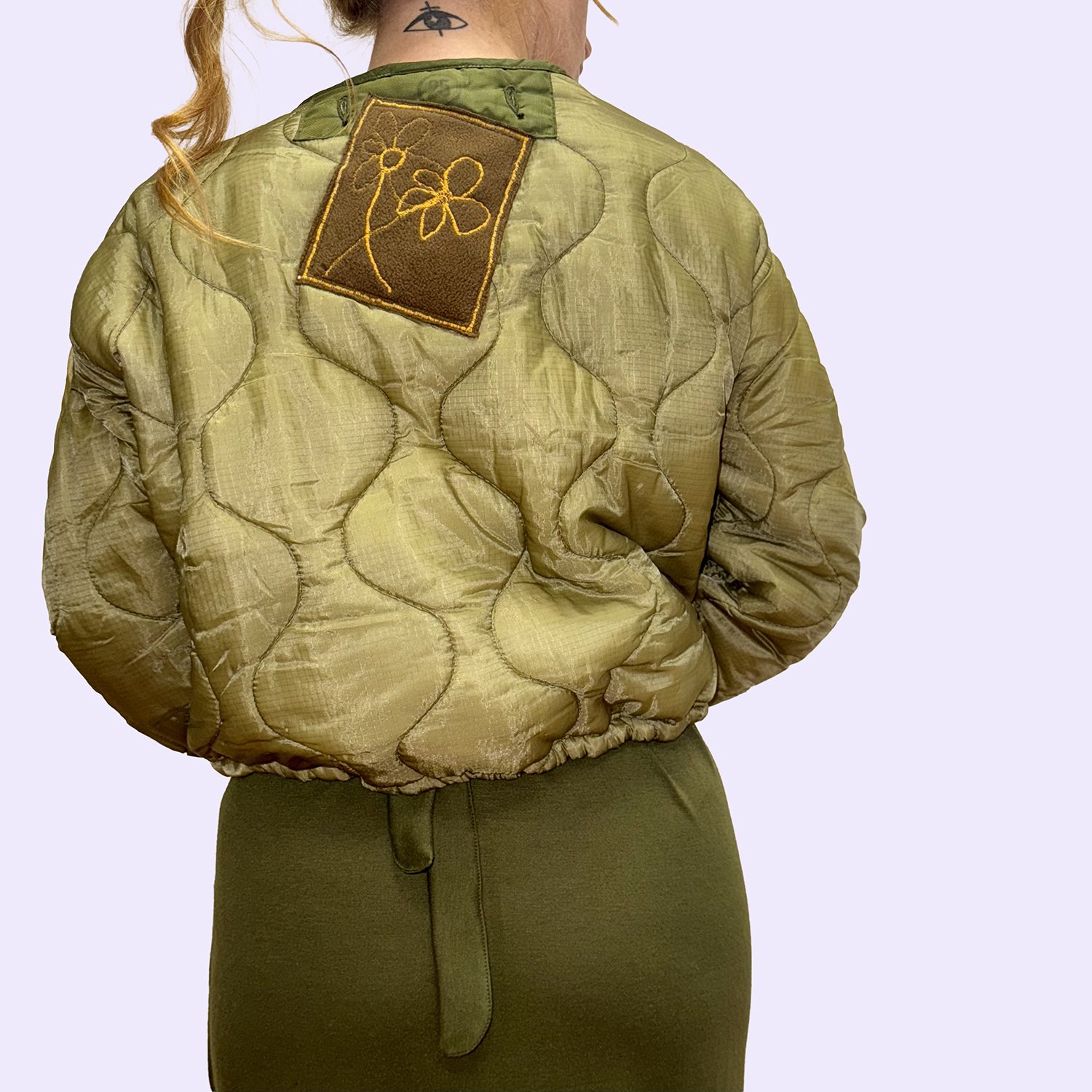 Femme portant une veste matelassée kaki avec un patch de broderie florale sur l'épaule gauche.