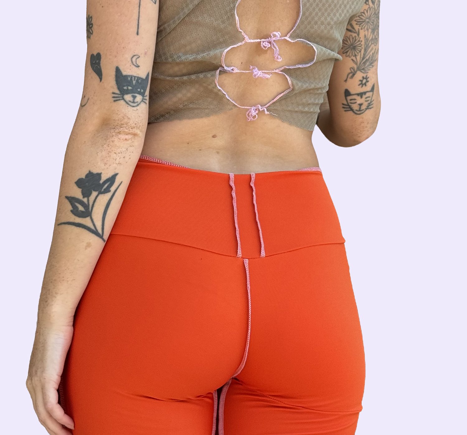 Une femme portant un haut beige dédoublé avec des liens roses serrés et un pantalon orange à couture rose, mettant en valeur ses tatouages sur les bras et l'abdomen.