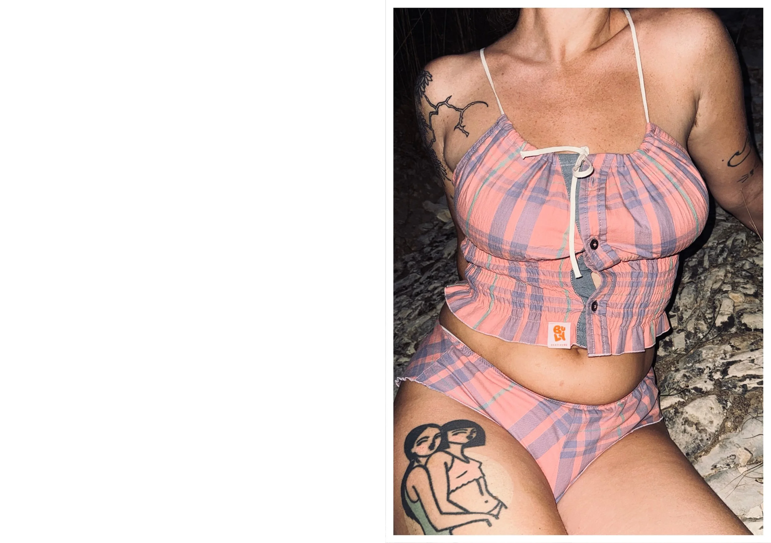 Une femme portant un haut à carreaux rose et une culotte assortie, assise sur des rochers, avec plusieurs tatouages visibles sur ses bras et sa cuisse.