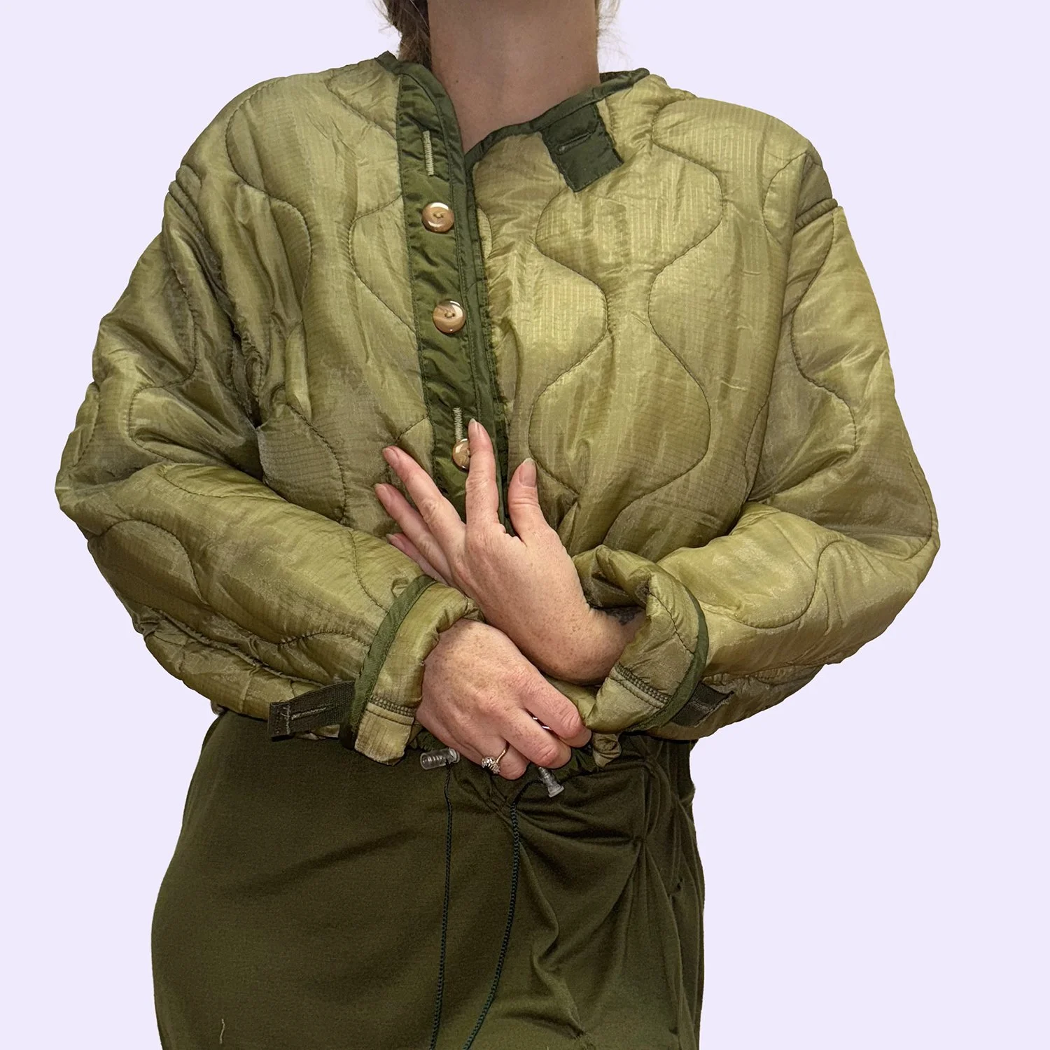 Une personne portant un manteau bouclé vert olive et un pantalon vert foncé, avec ses bras croisés et une main touchant l'autre.