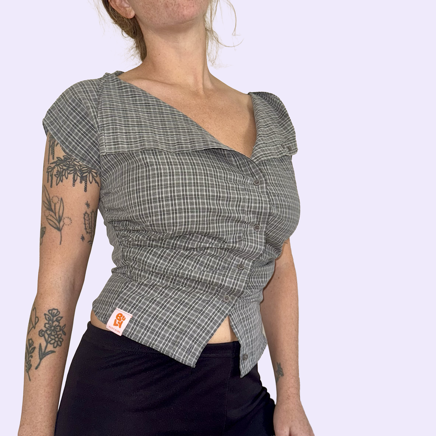 Une femme portant une chemise à carreaux gris, avec des tatouages sur le bras, devant un fond violet clair.
