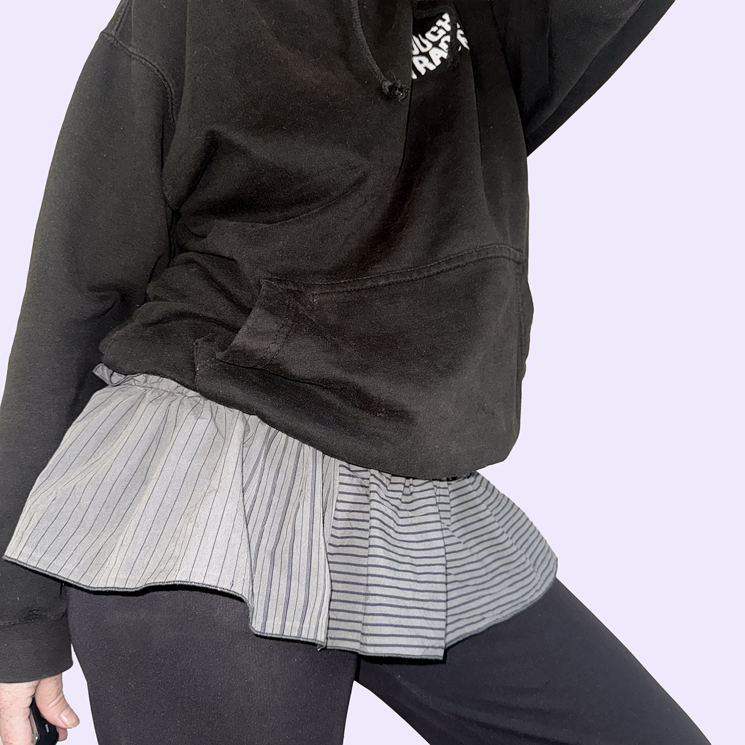 Personne portant un pull noir, une jupe plissée à rayures et un pantalon noir, fond violet clair.