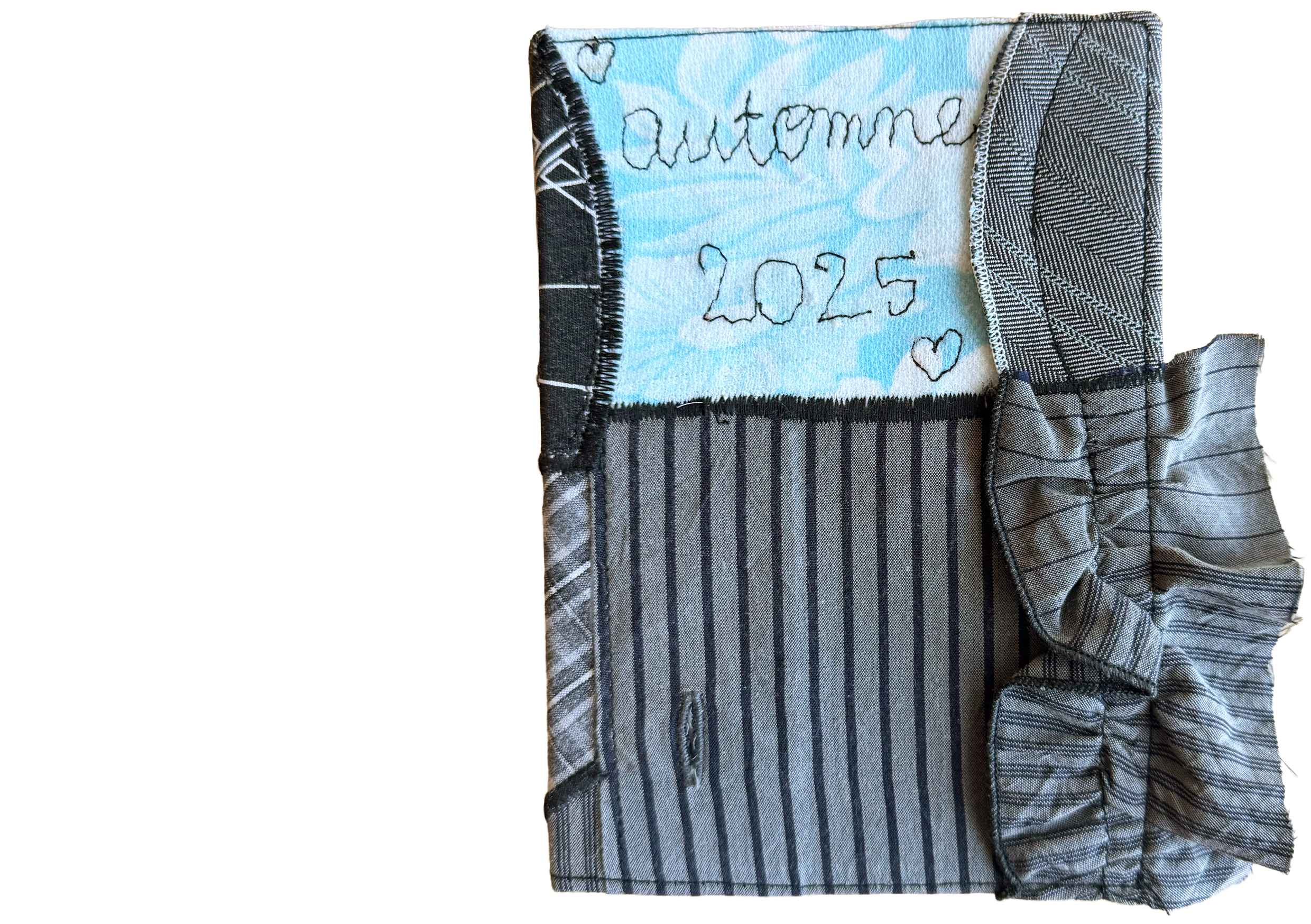 Un carnet avec une couverture en tissu rayé gris et un ruban en tissu également rayé. La page à l'intérieur est bleue avec écriture en fil noir indiquant 'automne 2025' et deux petits cœurs.