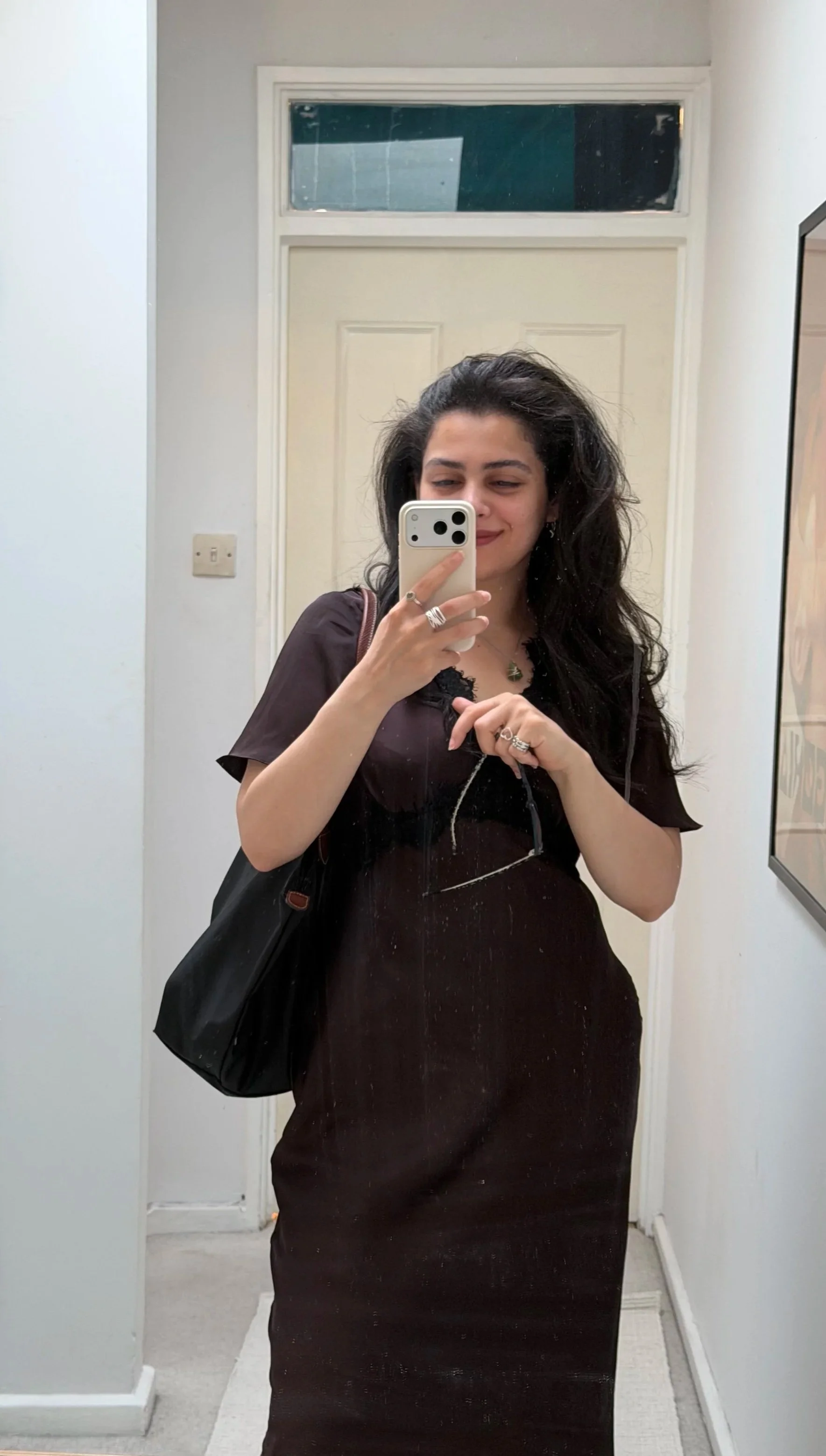 A woman taking a mirror selfie in a small room or hallway, holding a smartphone in her right hand, wearing a black dress and carrying a black purse on her left shoulder, with long dark hair and jewelry, smiling slightly.
