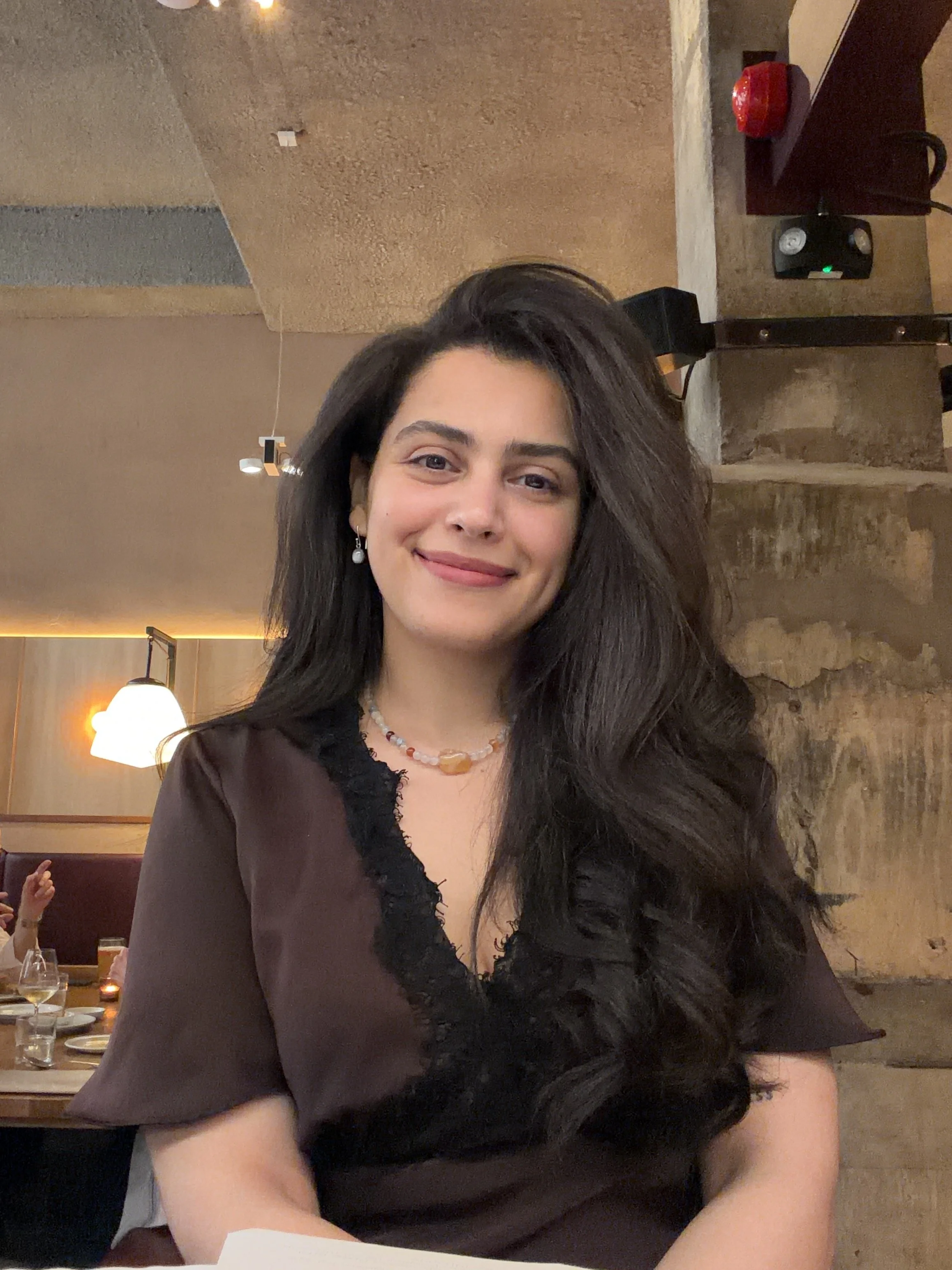 A woman with long, dark hair and earrings smiling at a restaurant or cafe with warm lighting, textured walls, and tables set with glasses and plates in the background.