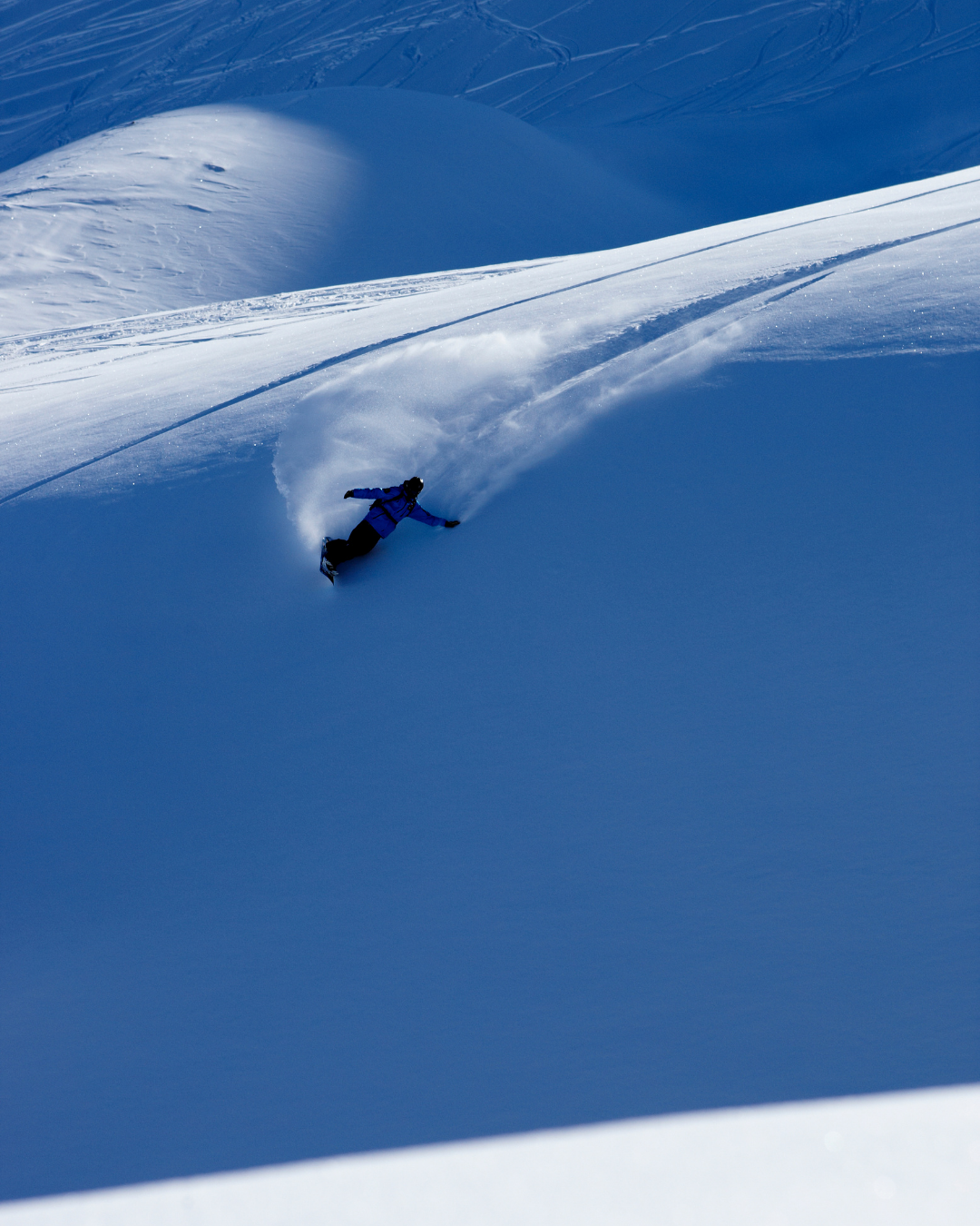 A skier in blue gear carving down a snowy mountain, kicking up a spray of snow behind them.
