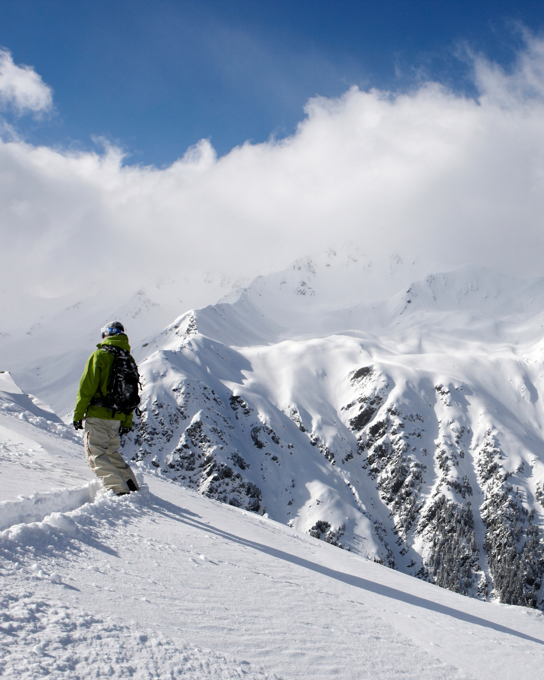 A person dressed in winter gear, including a green jacket, beige pants, and a backpack, standing on snowy mountain terrain with snow-covered peaks and a blue sky with clouds in the background.