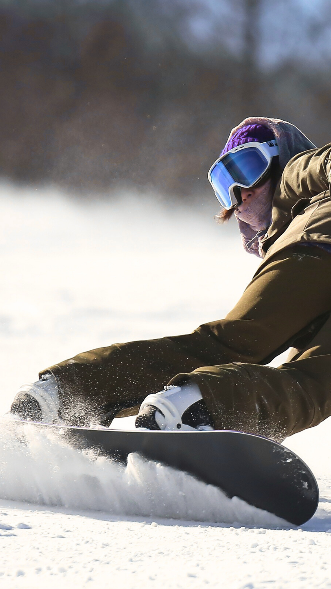Person snowboarding on snow-covered terrain, wearing goggles and winter clothing, with trees and cloudy sky in background.