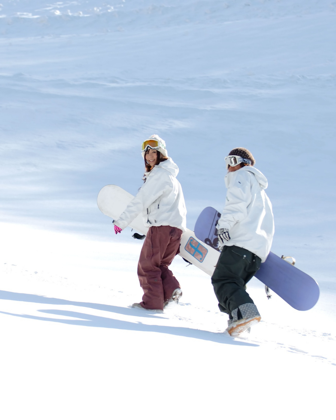 Two people wearing snow gear and helmets holding snowboards, walking on snow-covered slope with blue sky in background.
