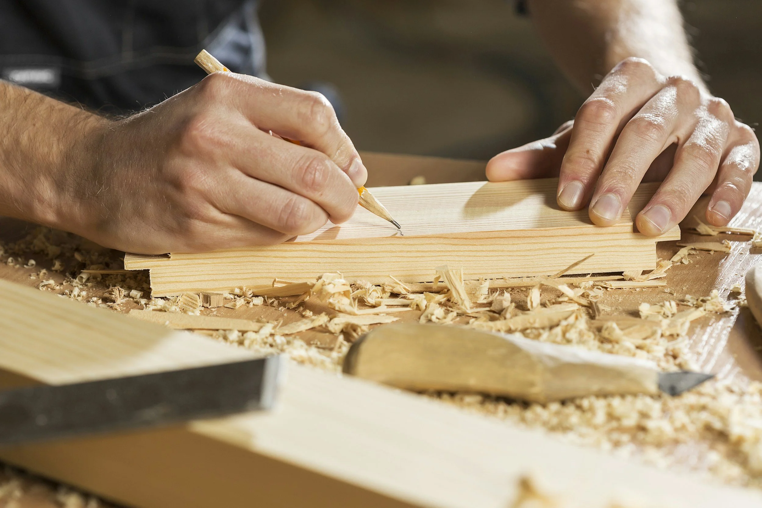 Close-up of hands carving a piece of wood with a pencil on a woodworking table, surrounded by wood shavings and woodworking tools.