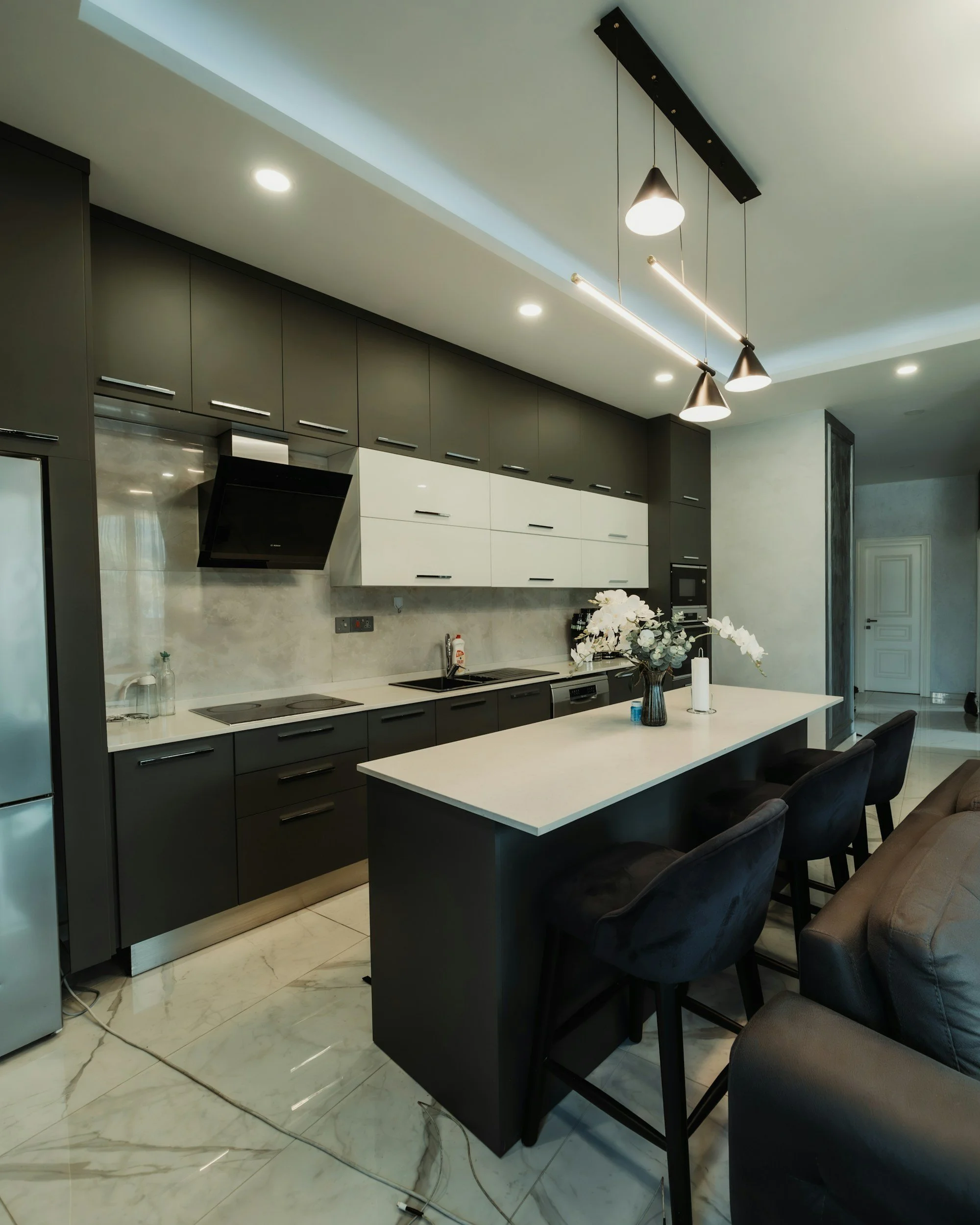 Modern kitchen with black and white cabinets, marble floor, kitchen island with black chairs, and contemporary lighting fixtures.