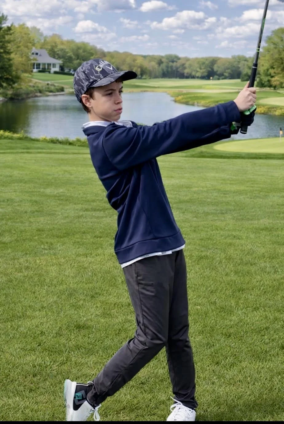 A boy wearing a baseball cap, navy sweatshirt, and black pants swings a golf club on a golf course near a pond and trees.