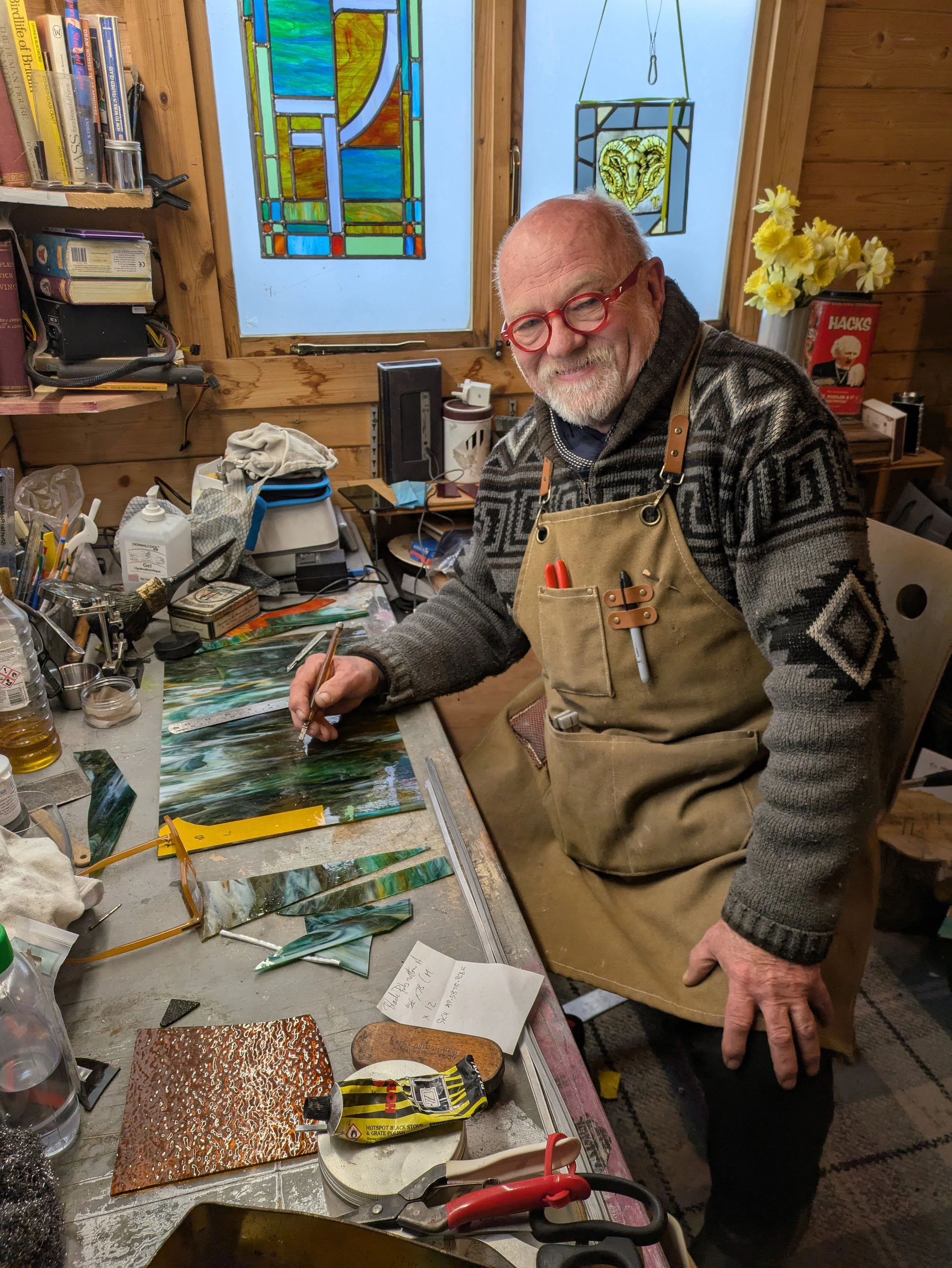 An elderly man with red glasses and a gray beard is working on a stained glass project inside a cozy wood-paneled workshop. He is seated at a cluttered worktable with various tools and colorful glass pieces, smiling as he works.
