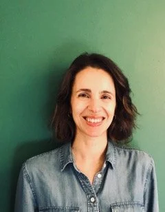 A woman with shoulder-length dark hair smiling, wearing a denim shirt, standing in front of a green background.