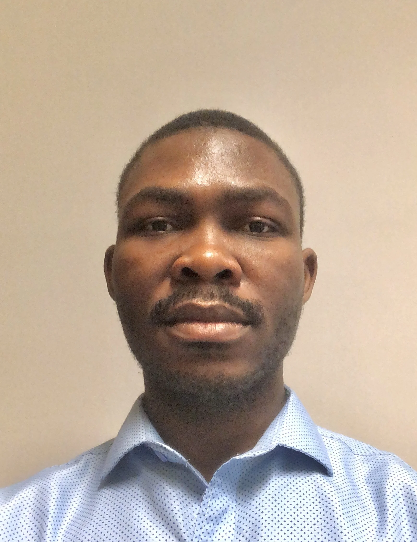 Portrait of a man with short hair and a beard, wearing a light blue button-up shirt, standing against a plain beige wall.