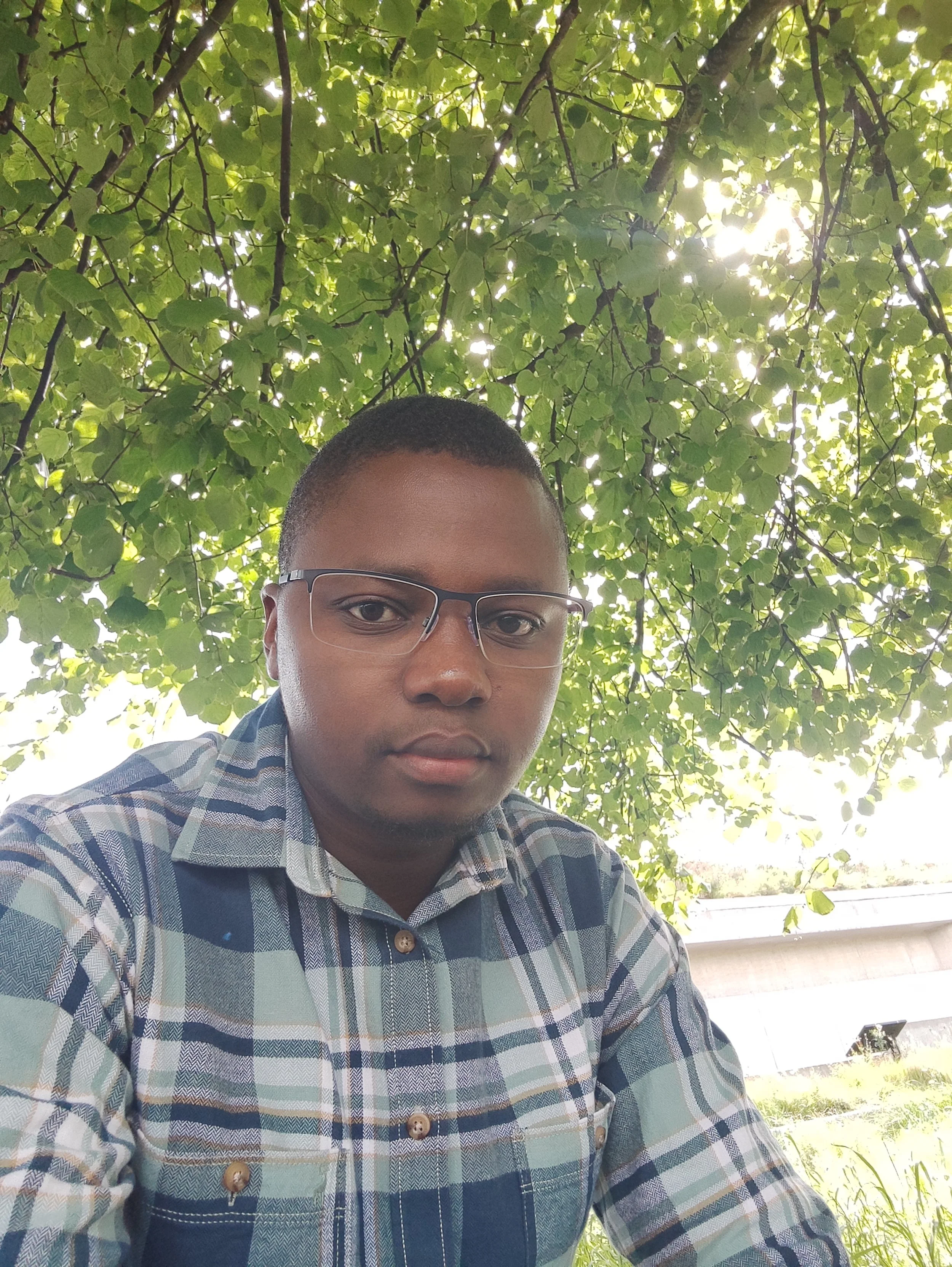 A young man wearing glasses and a plaid shirt, sitting outdoors under a tree with dense green leaves, sunlight filtering through.