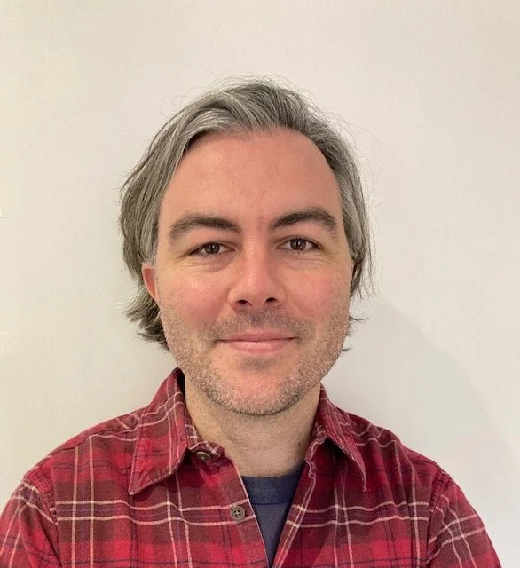 A man with light skin, gray hair, and a slight beard, wearing a red plaid shirt, smiling slightly in front of a plain white wall.
