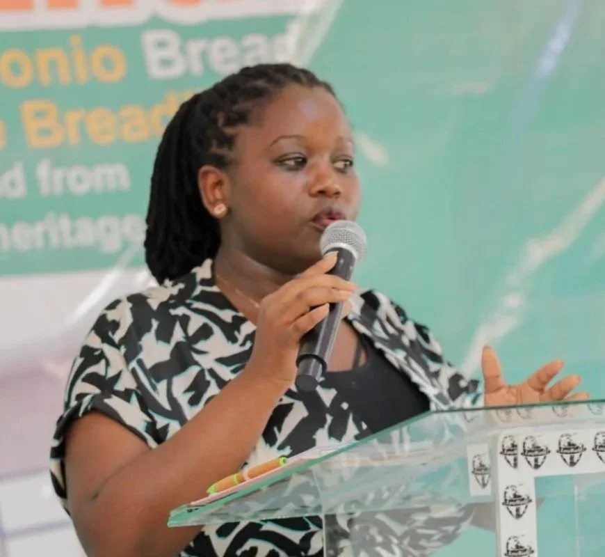 A woman with dark braided hair speaking into a microphone at a podium, wearing a black and white patterned shirt, with a blurred green background.