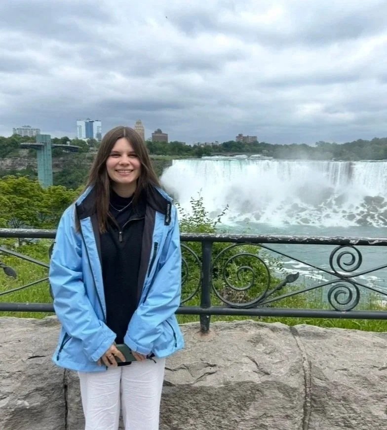 A young woman in a light blue jacket standing in front of Niagara Falls with a cityscape in the background.