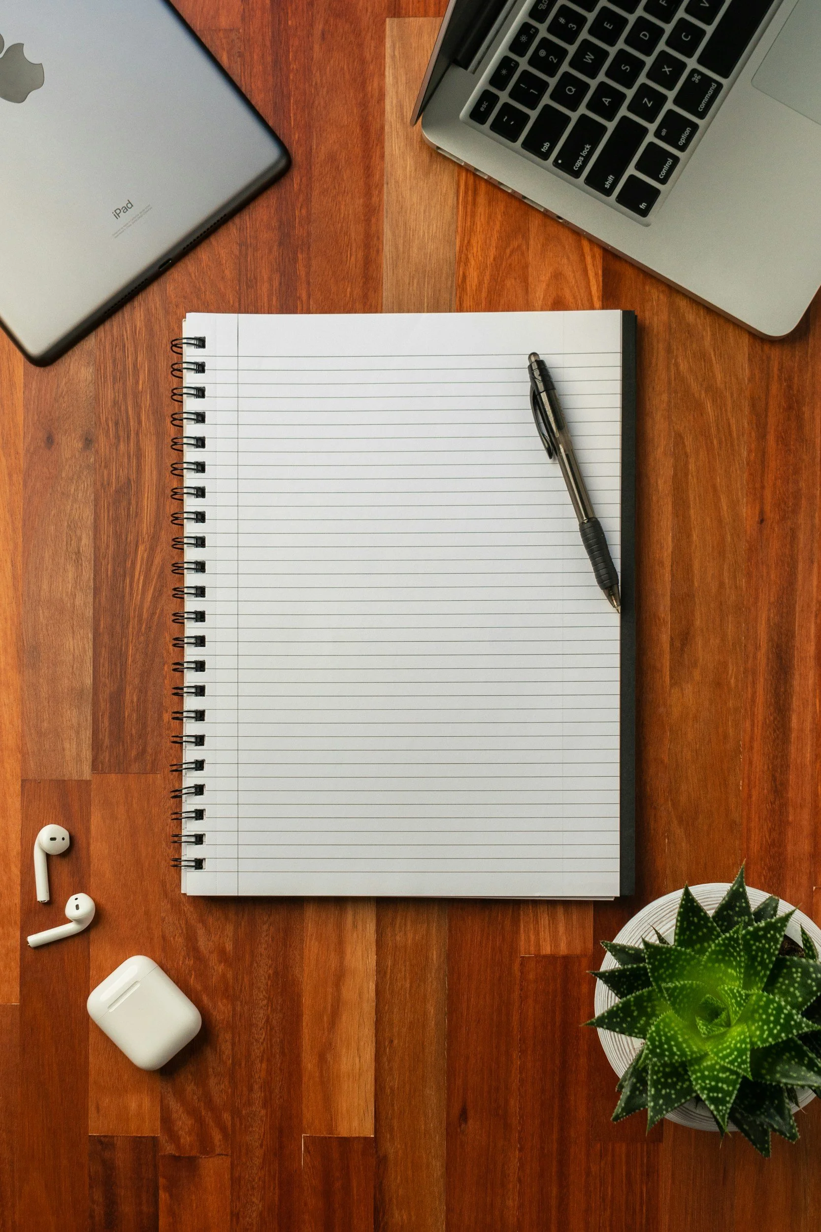 Top-down view of a wooden desk with an open spiral notebook, a pen, a pair of wireless earbuds, a charging case, a laptop keyboard, an iPad, and a potted succulent plant.