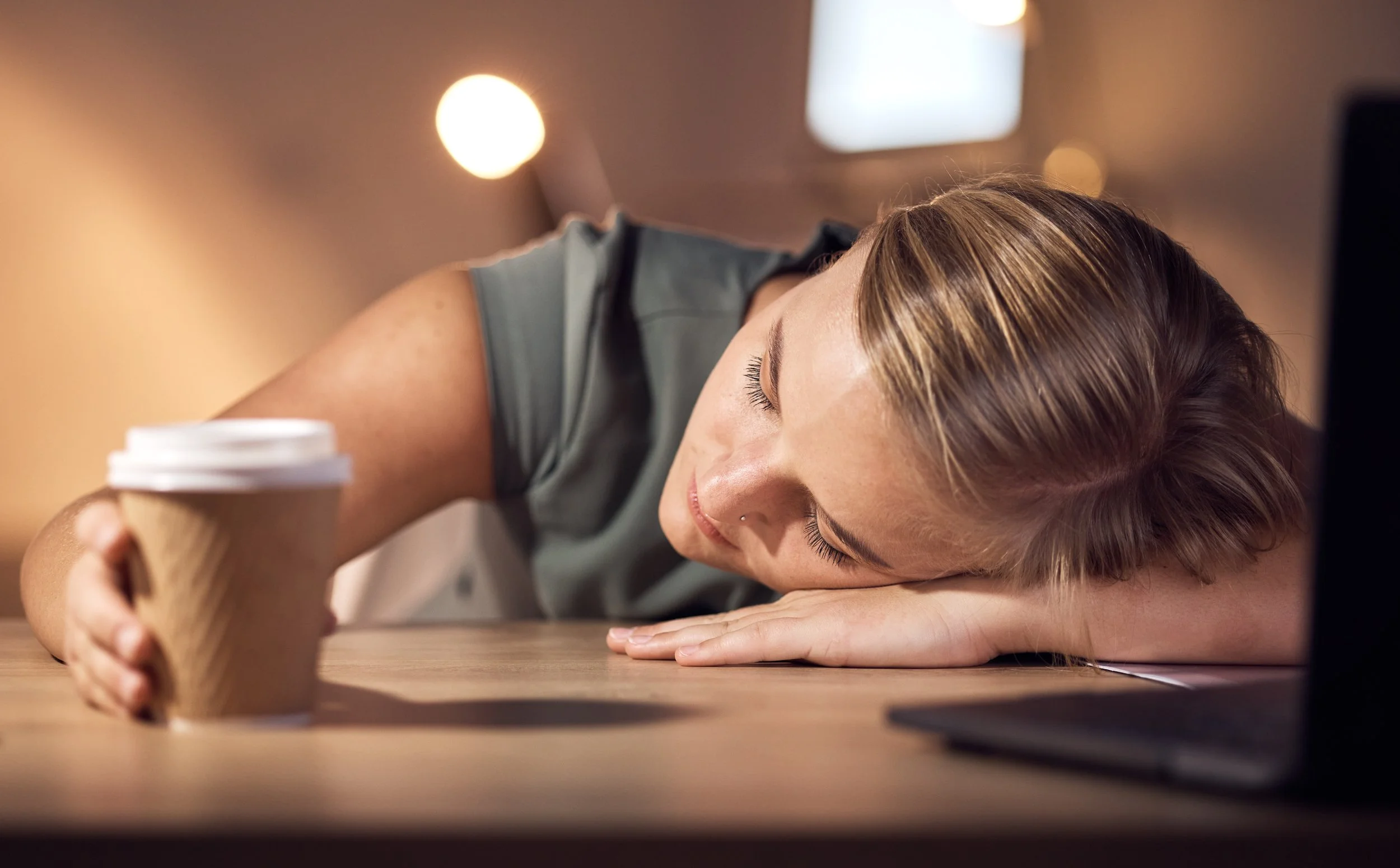 woman exhausted sleeping with a coffee in a hand in front of a computer