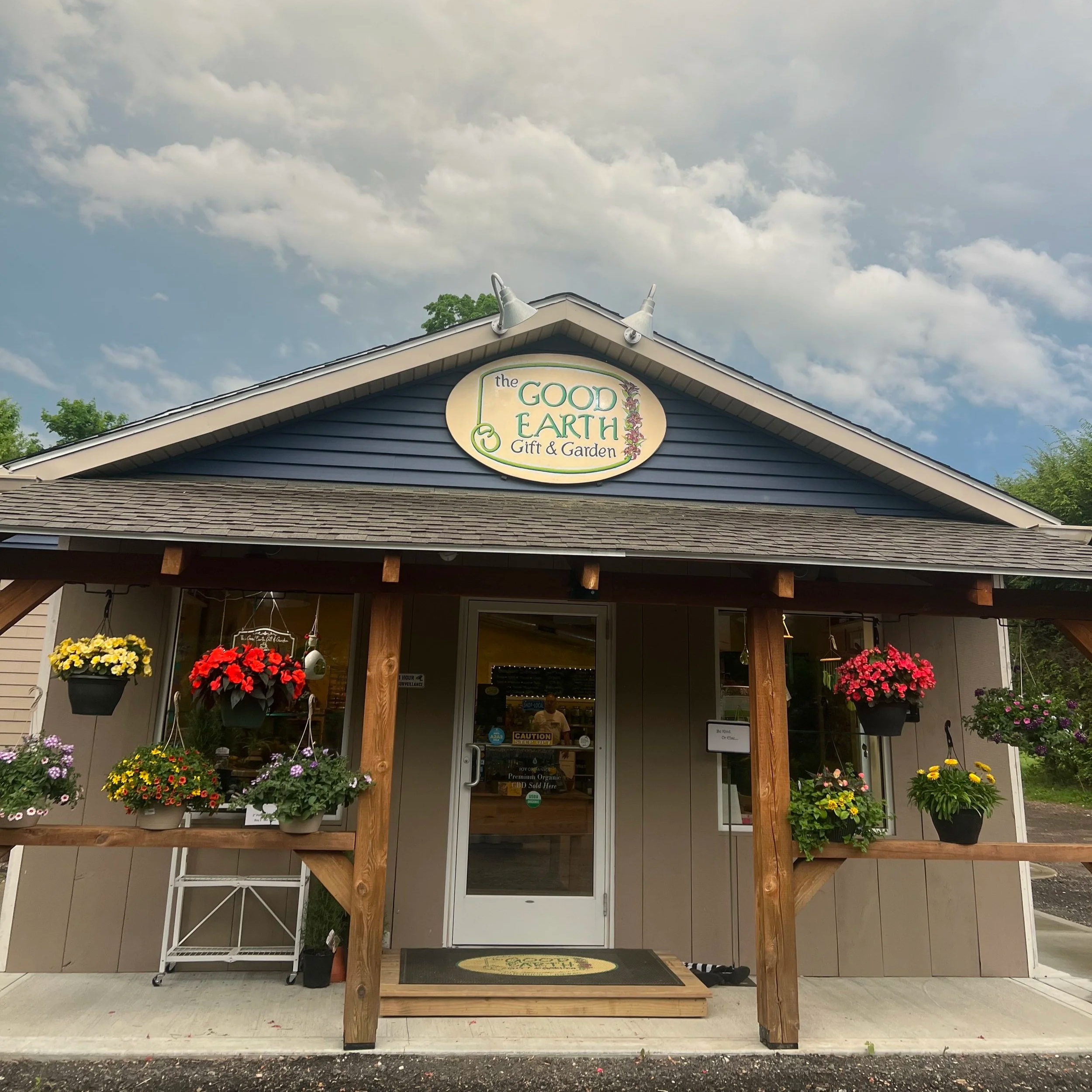 Front view of a building with a sign reading 'the GOOD EARTH GIFT & Garden'; the building has a blue exterior and a shingled roof, decorated with hanging flower baskets and potted plants, and wide glass doors with a small step in front.