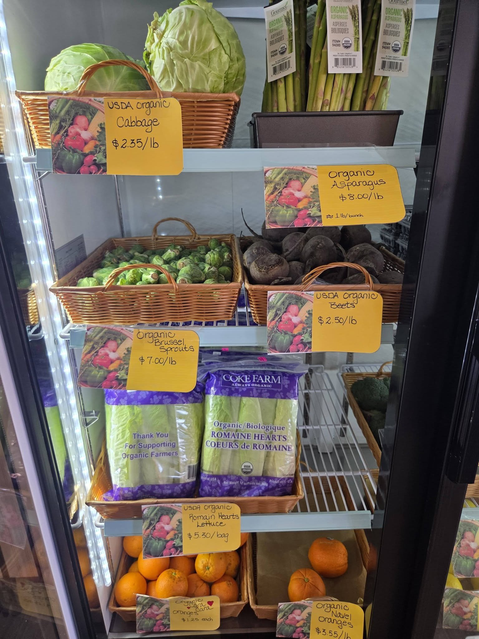 Display of organic vegetables and fruits in a refrigerated section at a store. Visible items include green cabbage, green Brussels sprouts, beets, and various oranges including Navel oranges, with handwritten price tags and labels.