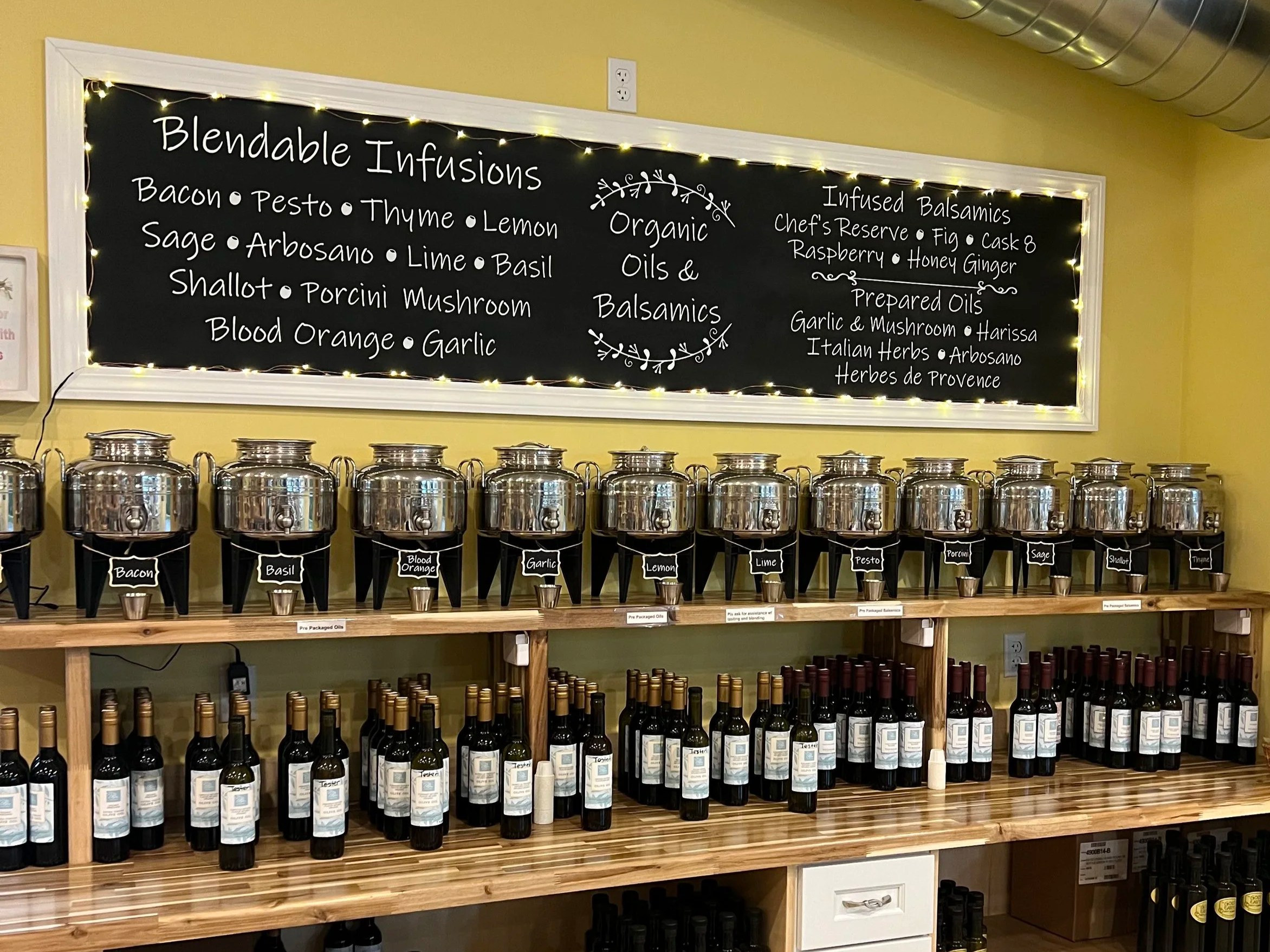 A display of various oils and balsamics in jars and bottles on wooden shelves, with a black signboard listing ingredients for flavored oils and balsamics, decorated with string lights.