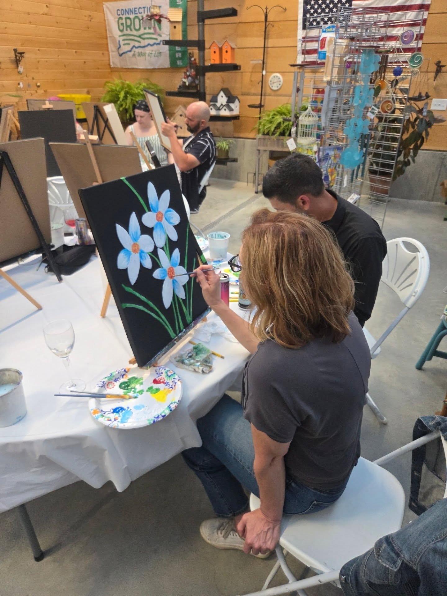 People painting a floral artwork on a canvas during an art class. One person is painting white flowers with orange centers on a black background.