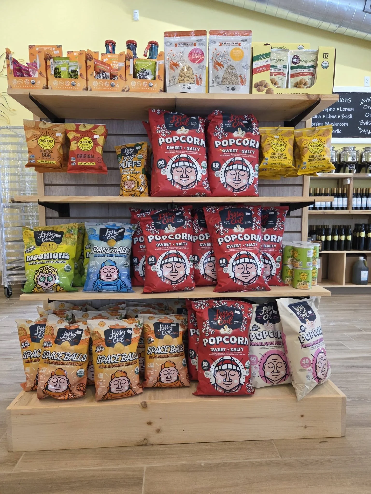 Display of various snack bags on wooden shelves in a store. The bags include popcorn, space balls, trail mix, and other organic snacks.