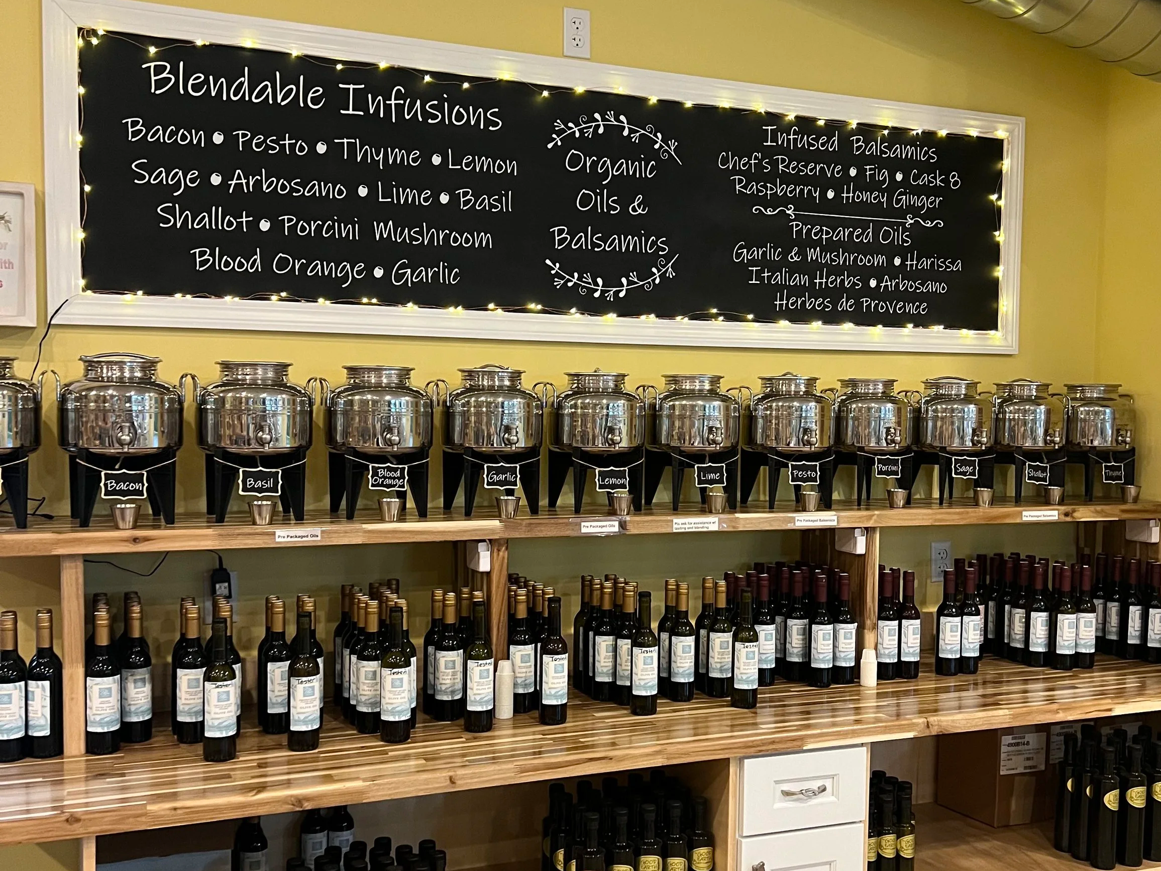 A chalkboard menu with the title 'Blendable Infusions' and lists ingredients like bacon, pesto, thyme, lemon, sage, arugula, lime, basil, shallot, porcini mushroom, blood orange, and garlic. The board also mentions 'Organic Oils & Balsamics' and 'Infused Balsamics' with flavors like fig, raspberry, honey ginger, garlic & mushroom, harissa, Italian herbs, arugula, and herbs de Provence. Below the chalkboard are metal containers labeled with various ingredients and bottles of wine on the shelves.