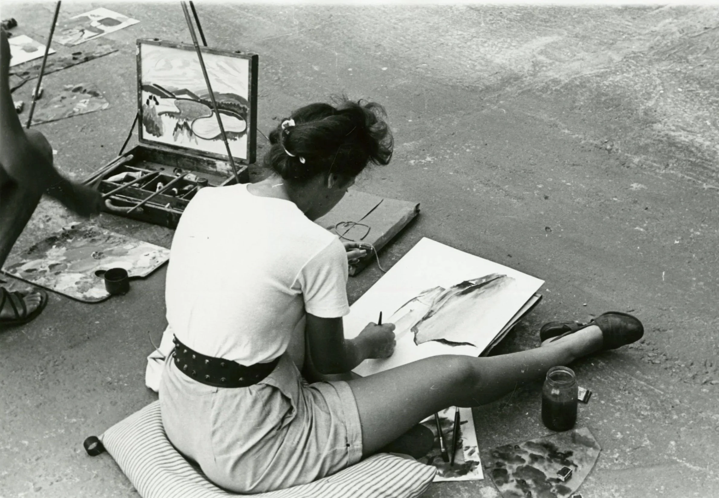 A person sitting on the ground and painting on paper, with art supplies and paintings around them on the street.