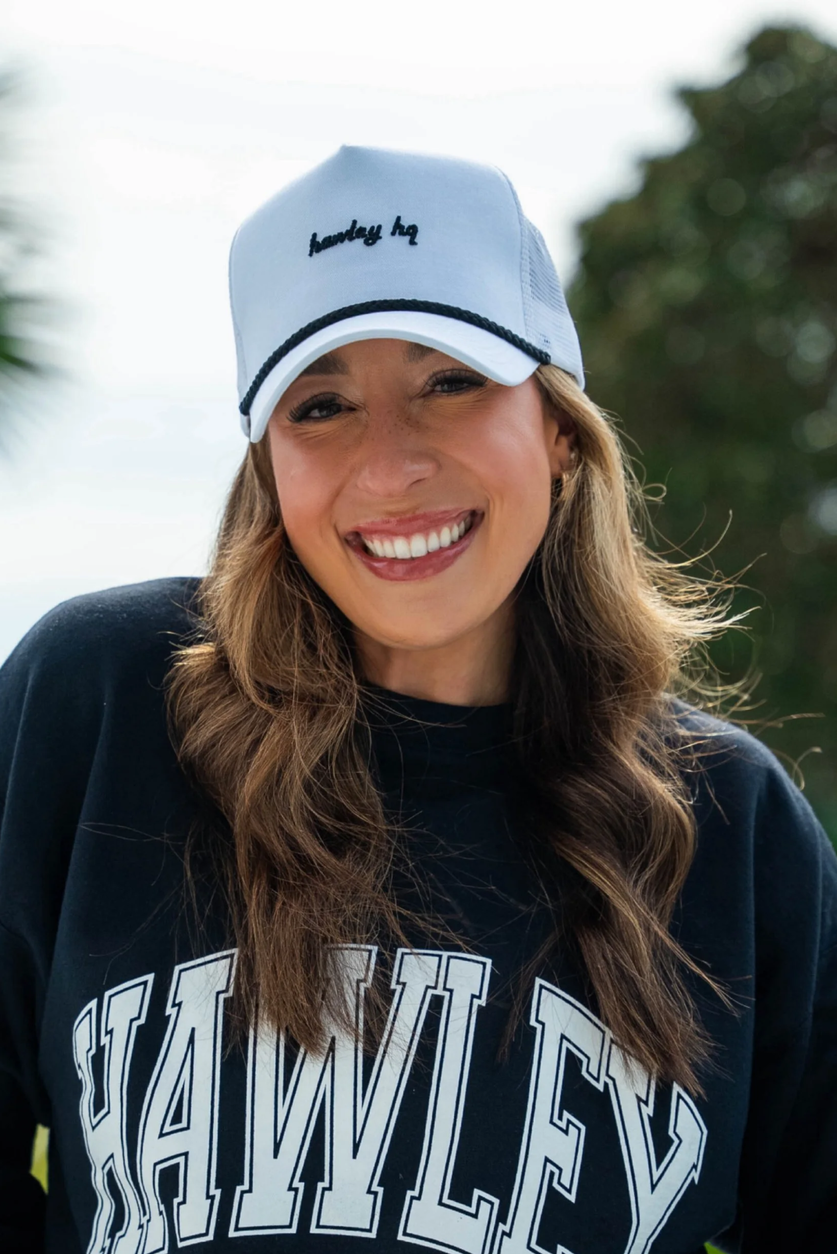 A woman wearing a white cap with black embroidery that reads 'sunday hug' and a dark sweatshirt with large white letters spelling 'HARVARD'.