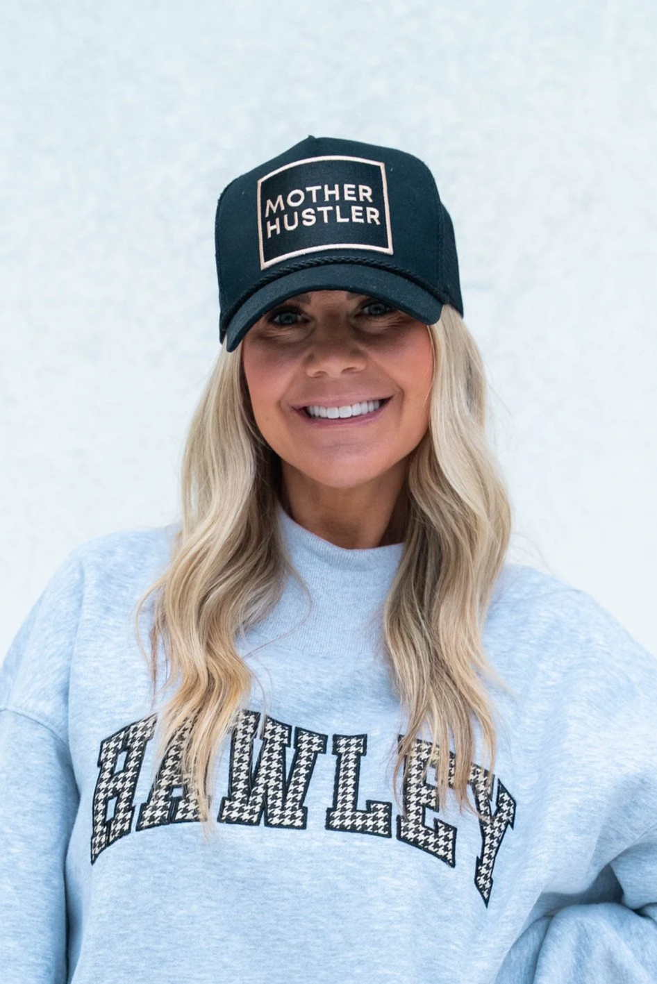 A woman with long blonde hair smiling, wearing a gray hoodie with 'BABY LXY' printed on it and a black cap that reads 'Mother Hustler'.