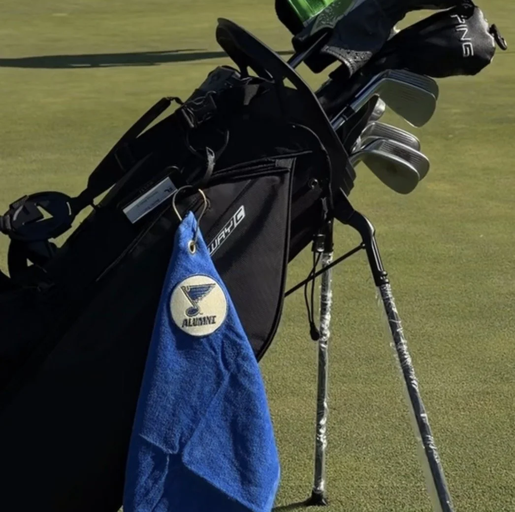 A golf bag with clubs and a blue custom branded towel hanging from it, located on a golf course with green grass.
