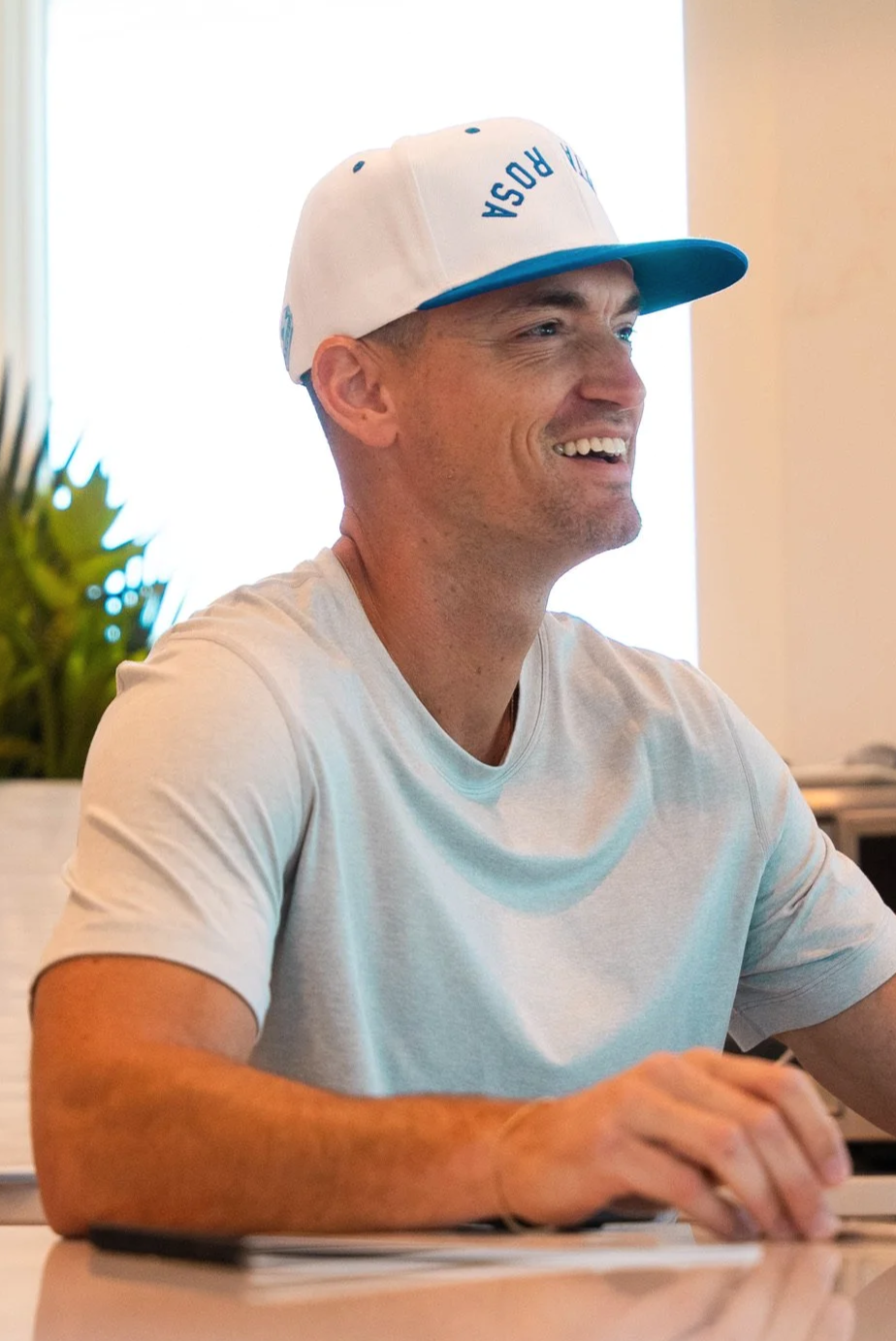 A man wearing a white cap with blue text and a white t-shirt, smiling and looking to the right while sitting at a table.