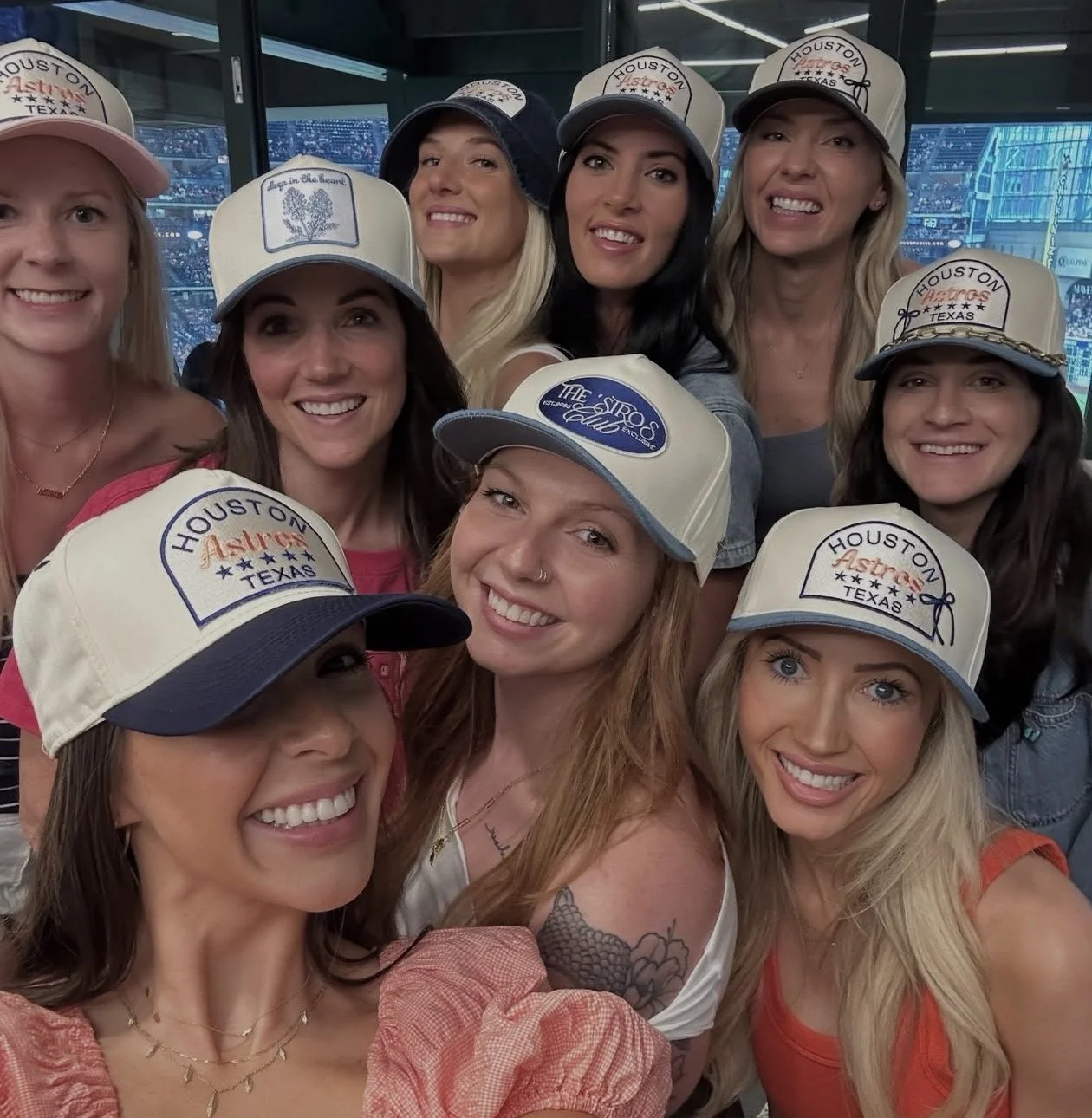 Group of ten women smiling and taking a selfie at a stadium, all wearing custom Houston Astros baseball caps.