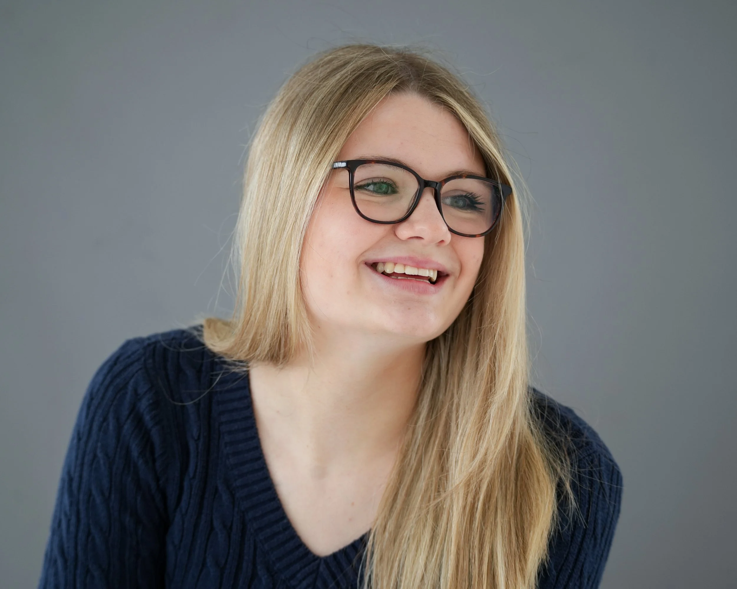 Close-up of a young woman with blonde hair and green eyes, wearing glasses and a navy blue sweater, smiling against a gray background.