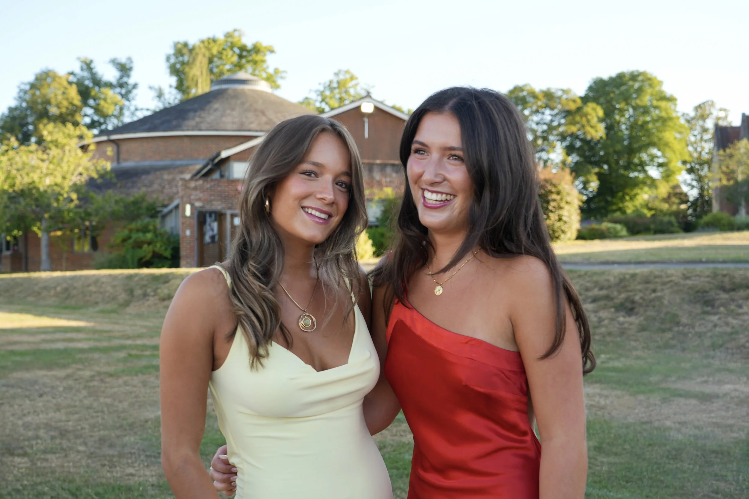 Two women standing outdoors in front of a house with trees, smiling and wearing dresses in cream and red colors.