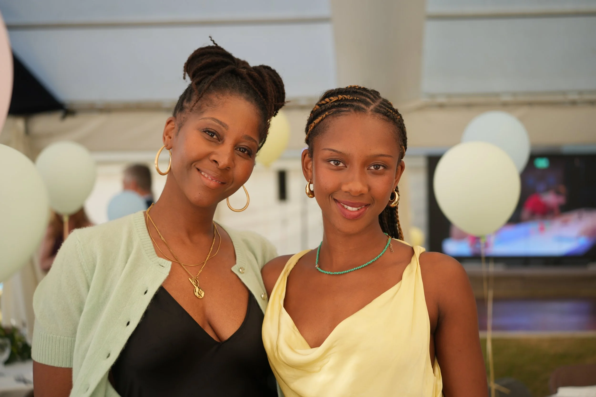 Two smiling women, one younger and one older, standing close together at an indoor event with balloons in the background.