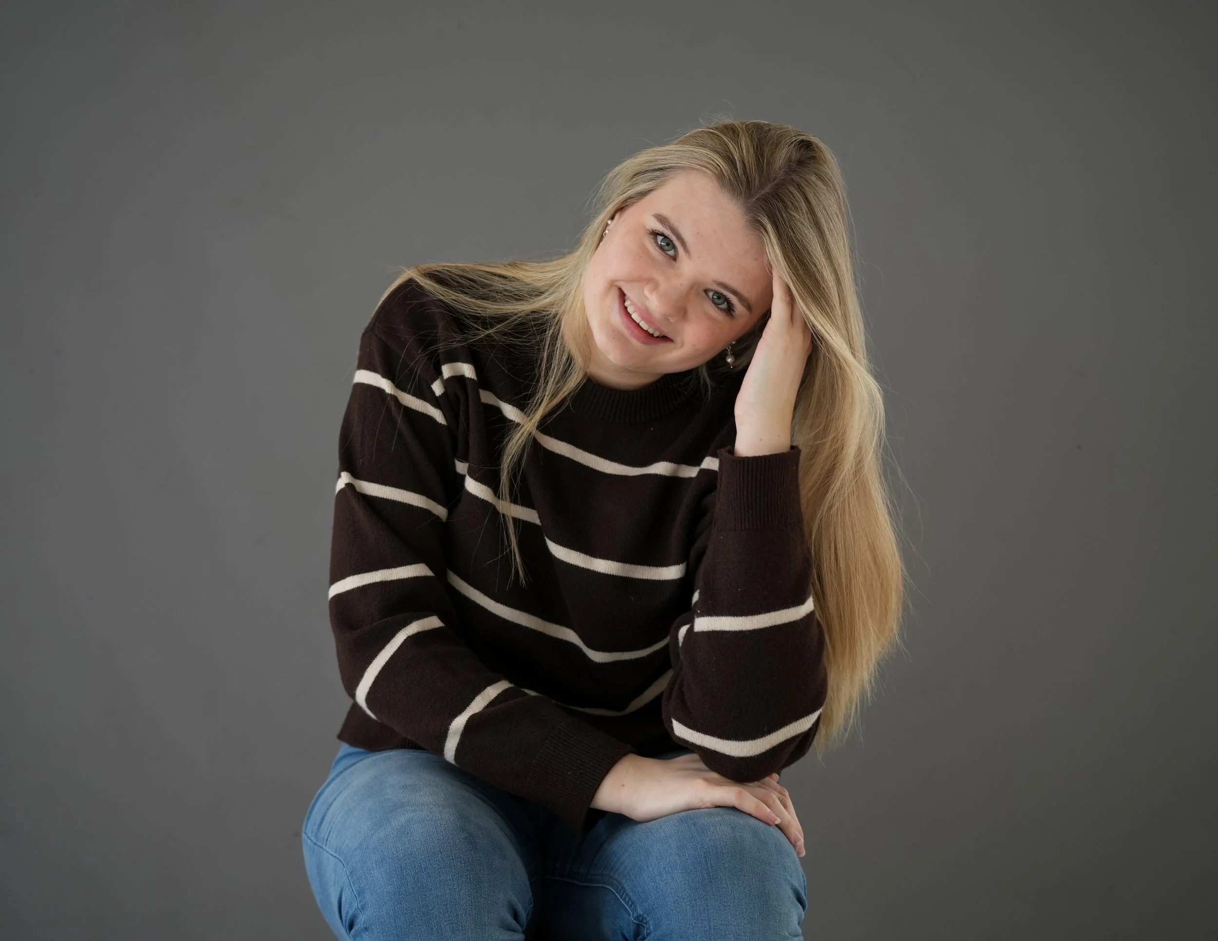 Young woman with blonde hair, blue eyes, smiling, wearing a black and white striped sweater and blue jeans, sitting against a gray background.