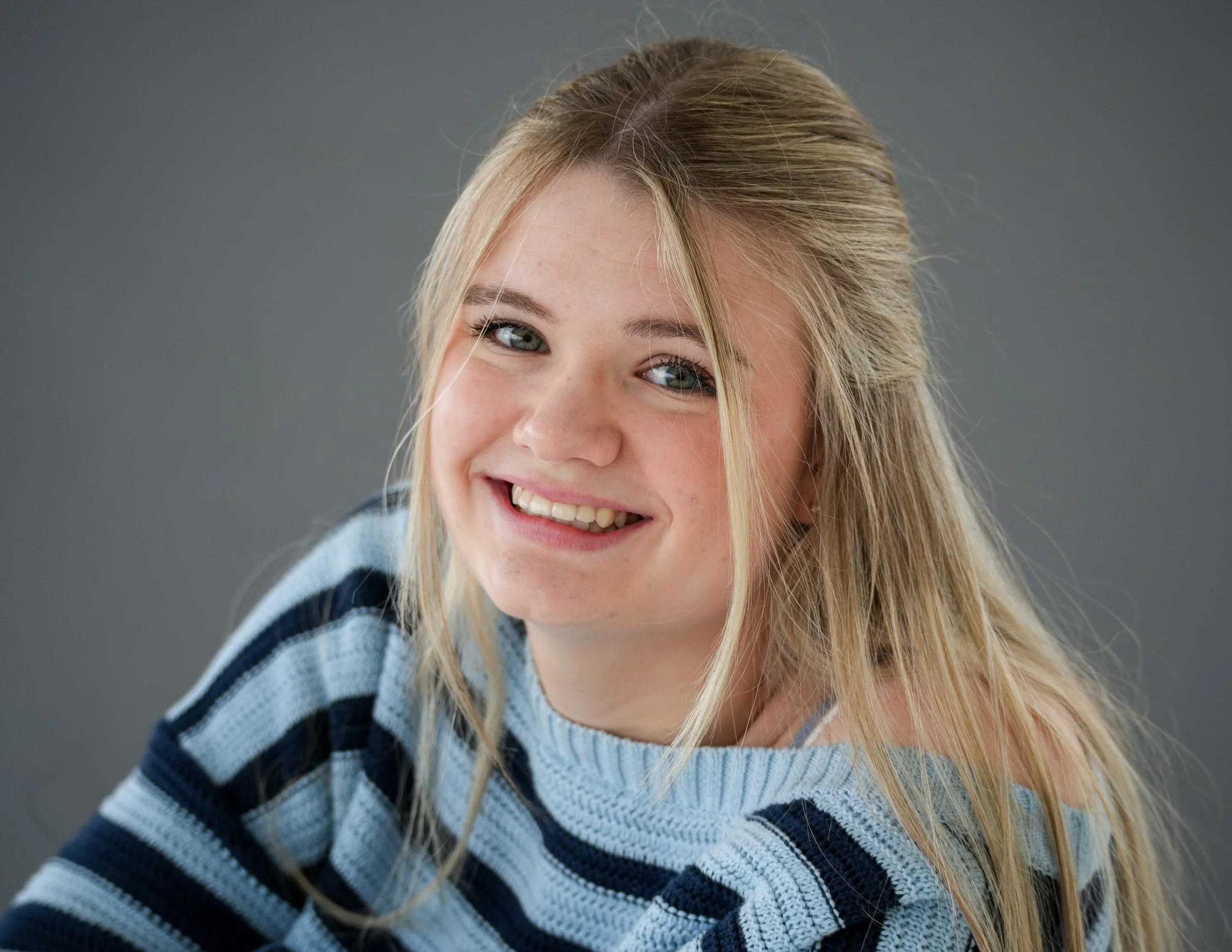 Close-up of a young girl with blonde hair, wearing a blue and white striped sweater, smiling in front of a gray background.