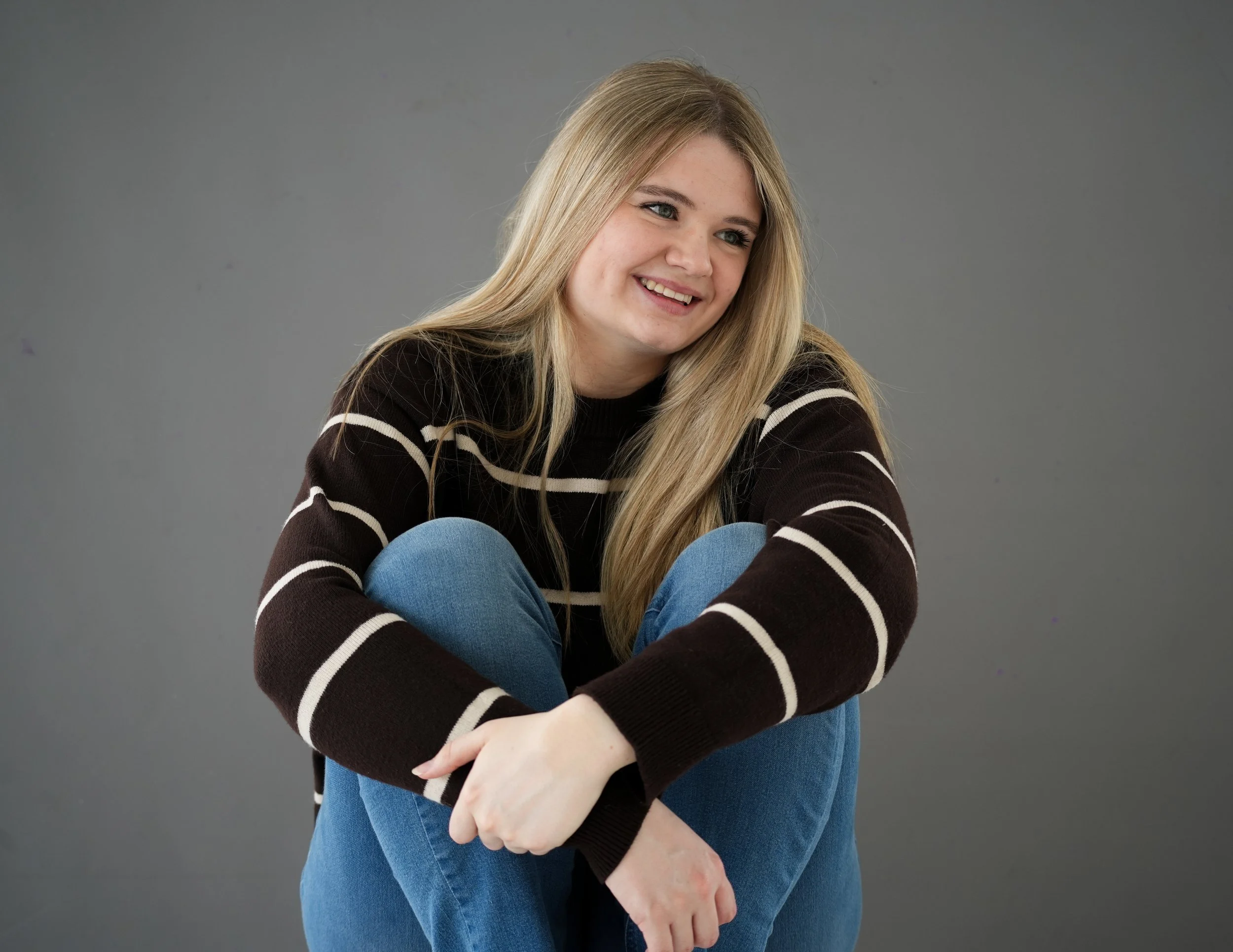 A young woman with long blonde hair, wearing a black sweater with white stripes and blue jeans, sitting against a gray background, smiling and looking to the right.