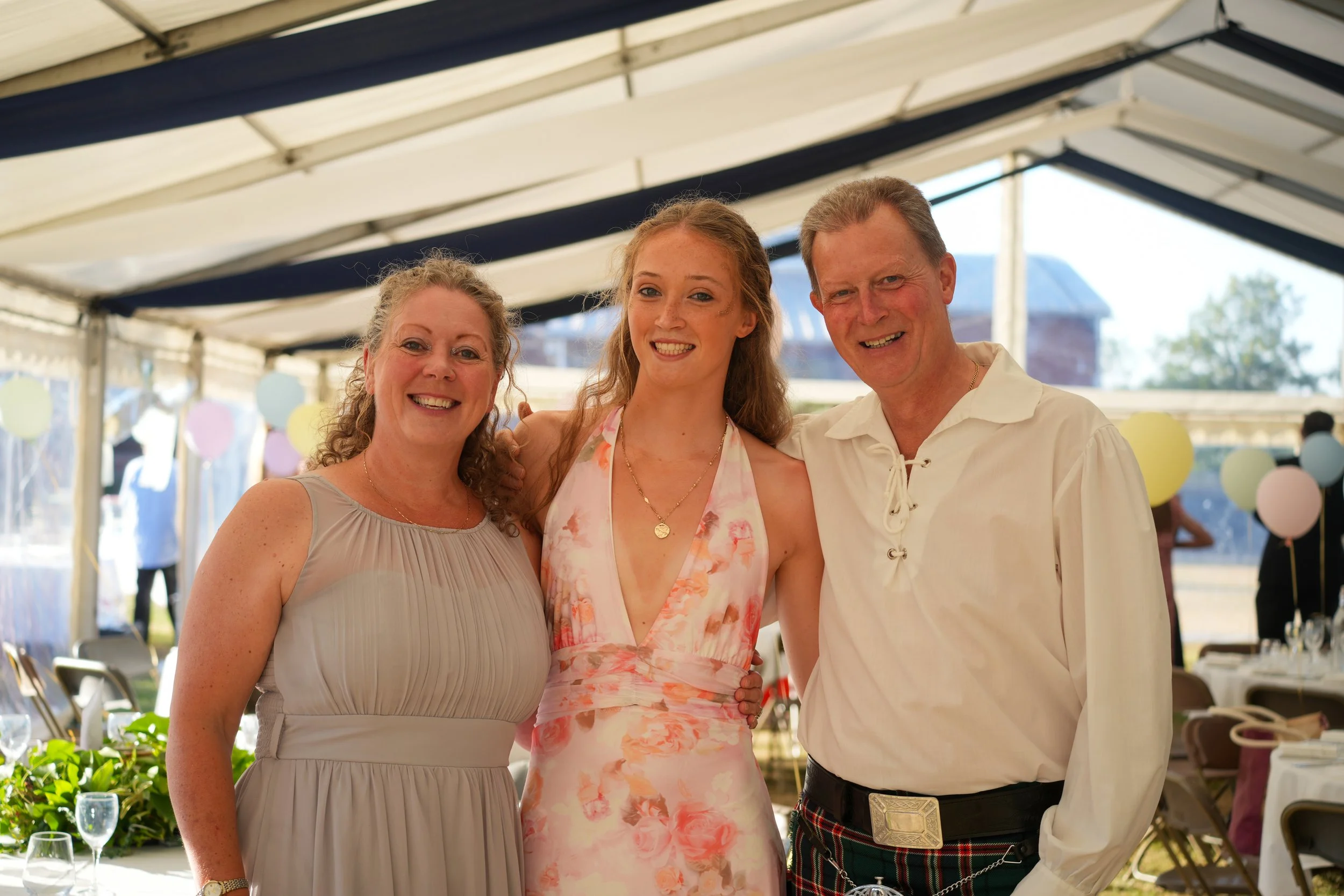 Three people smiling at a celebration under a white and blue venue tent, decorated with pastel balloons.