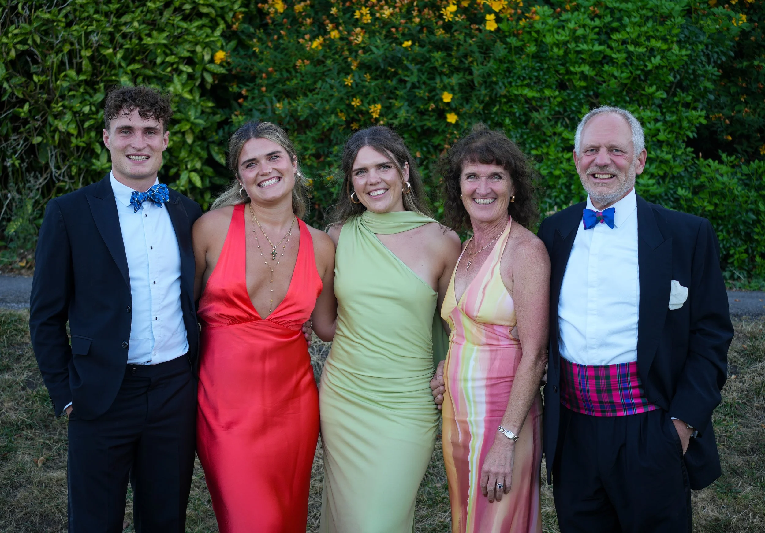 A family group photo of five people standing outdoors in front of green bushes with yellow flowers, smiling at the camera. The group includes two older adults and three younger adults or teenagers, dressed in semi-formal and colorful attire suitable 