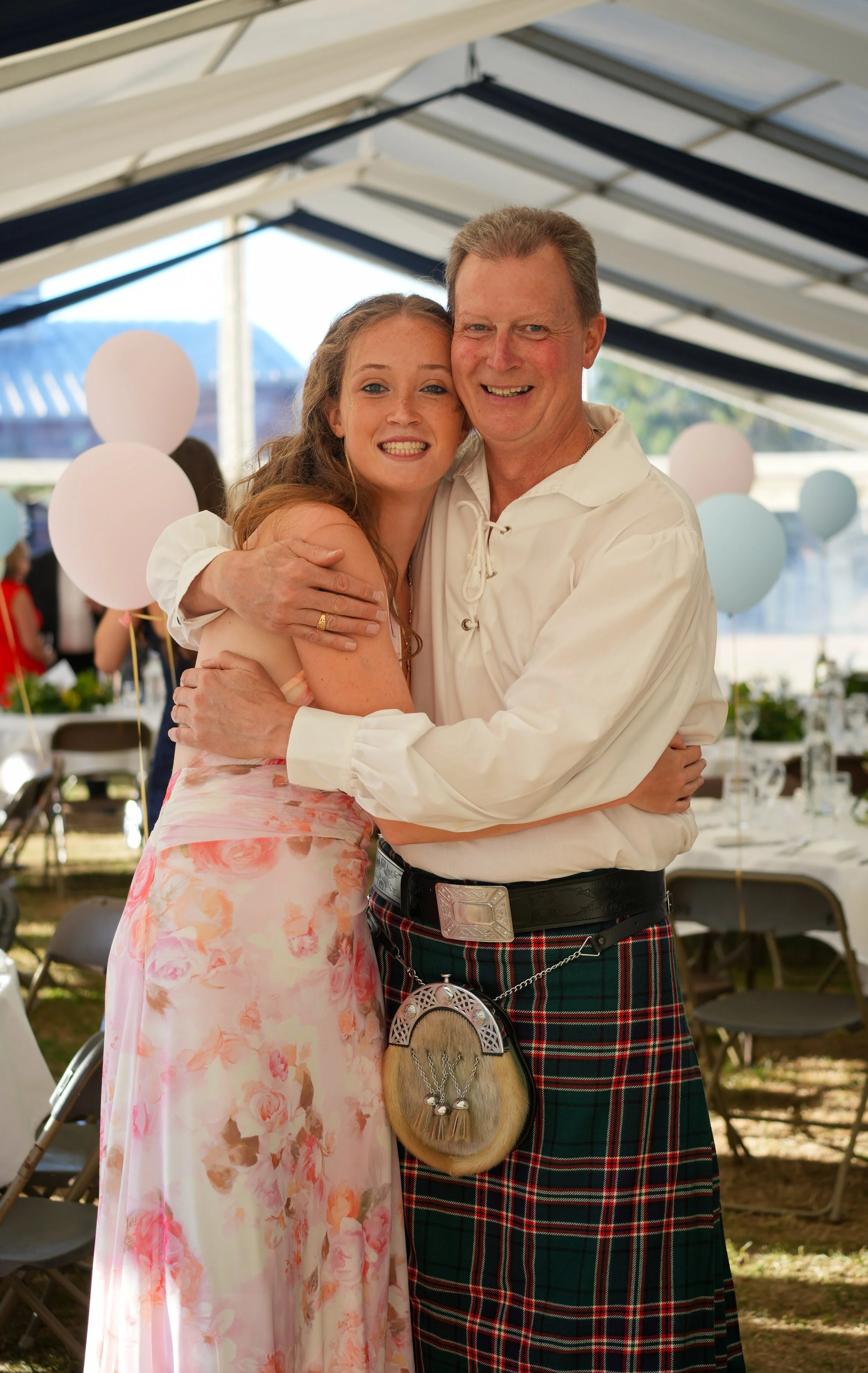 A young woman and an older man embracing and smiling at the camera at a daytime outdoor event under a tent, with balloons and tables in the background.