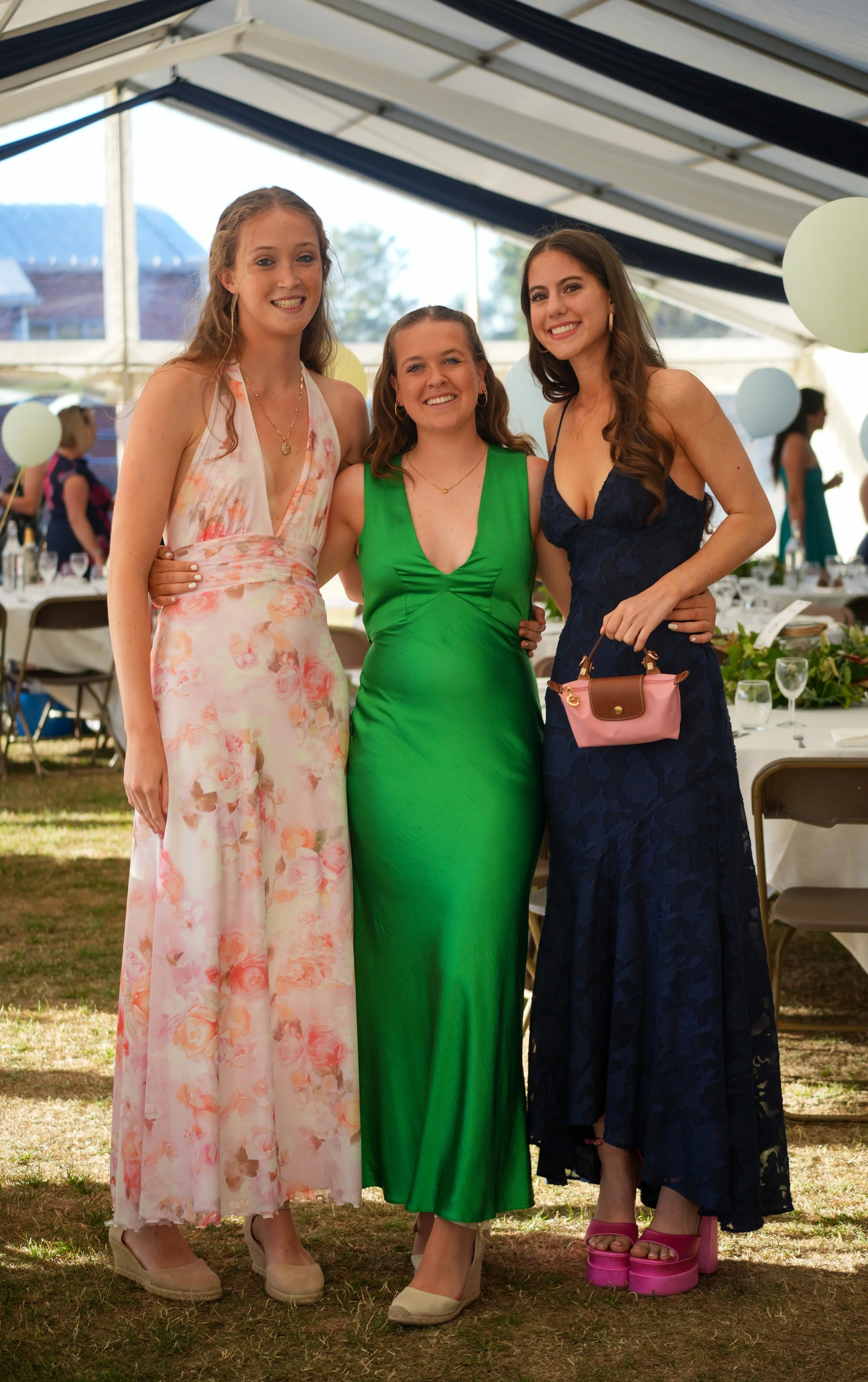 Three women standing inside a tent, smiling at the camera. They are dressed in colorful, elegant dresses and wearing high heels. There are tables with decorations and balloons in the background, indicating a celebration or event.